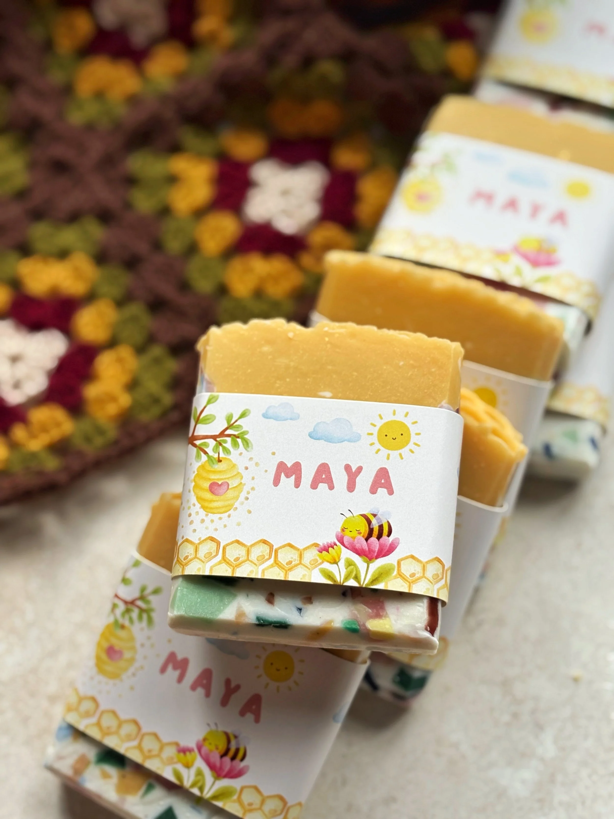 Bar of handmade soap with a colorful label featuring a bee, honey, flowers, and the name 'Maya' on a light background, with other soap bars and a crocheted blanket with floral patterns in the background.