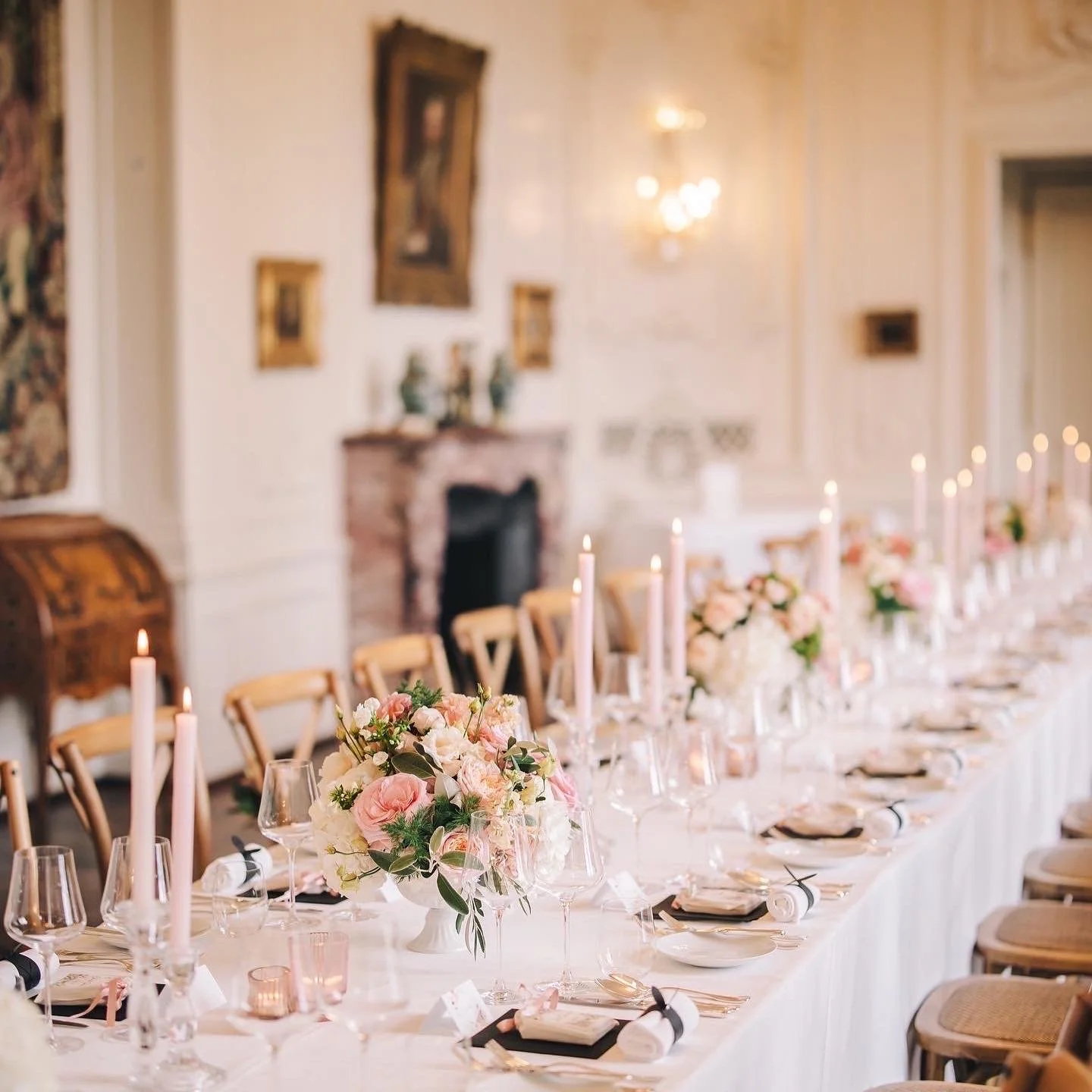 Elegant banquet table with pink and white floral centerpieces, pink candles, wine glasses, and place settings in a decorated room with artwork and a fireplace.