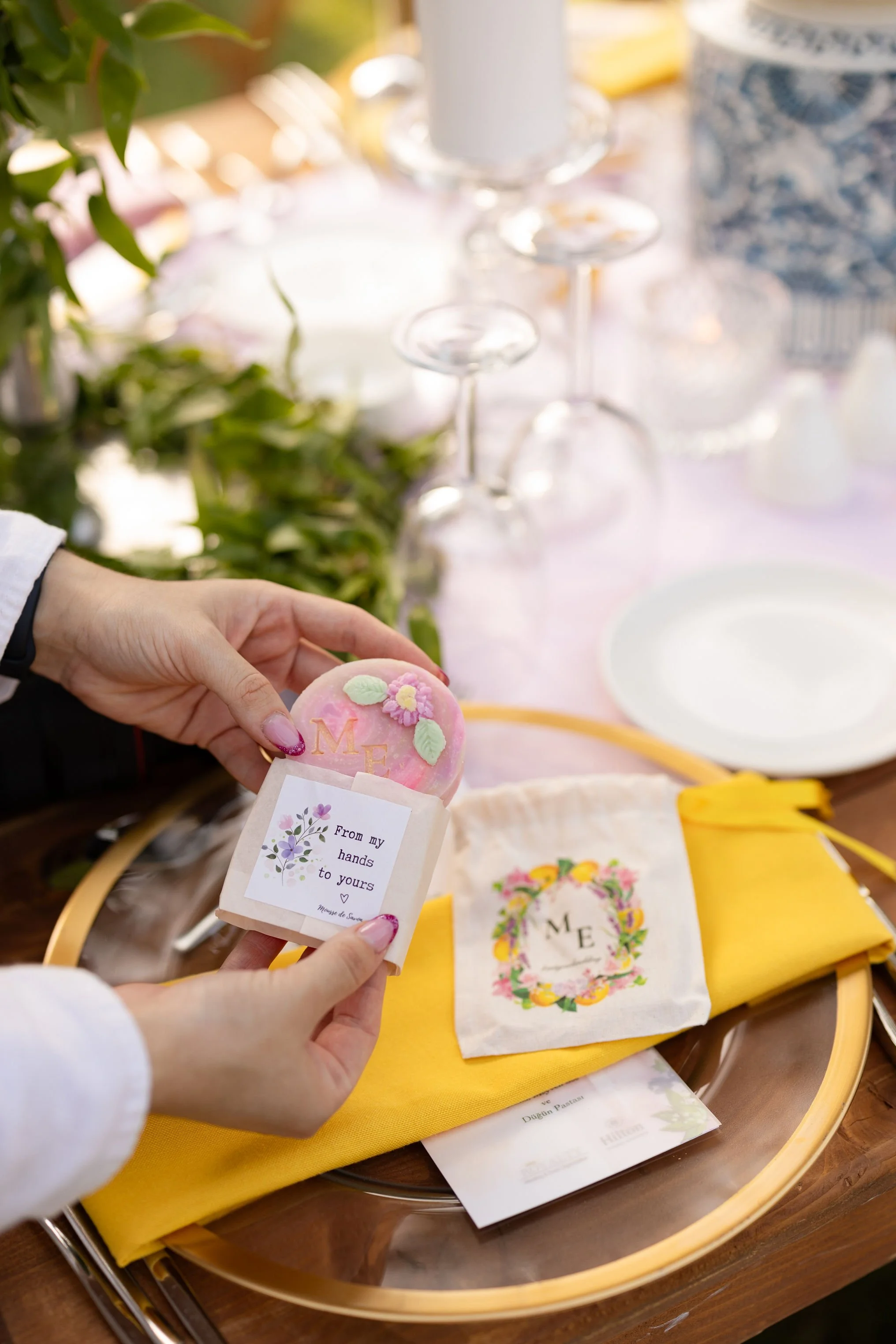 A person holding a decorated pink cookie with a letter 'M' and floral decorations, at a table set for a celebration with yellow and floral-themed napkins, empty plates, and glassware.