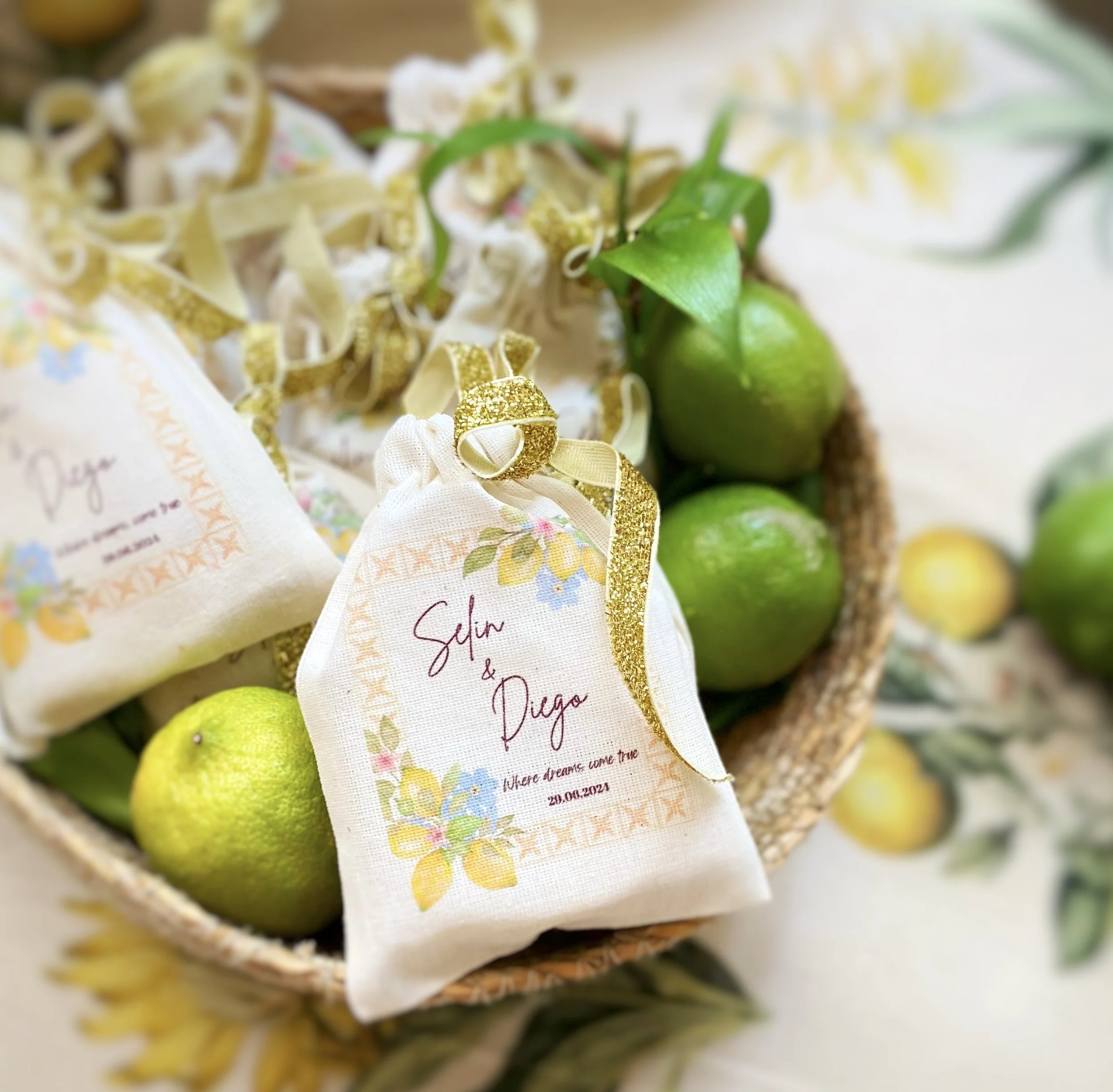 A basket of limes with small gift bags labeled 'Sijin & Diego' and 'Where dreams come true,' decorated with a gold ribbon, on a floral tablecloth.