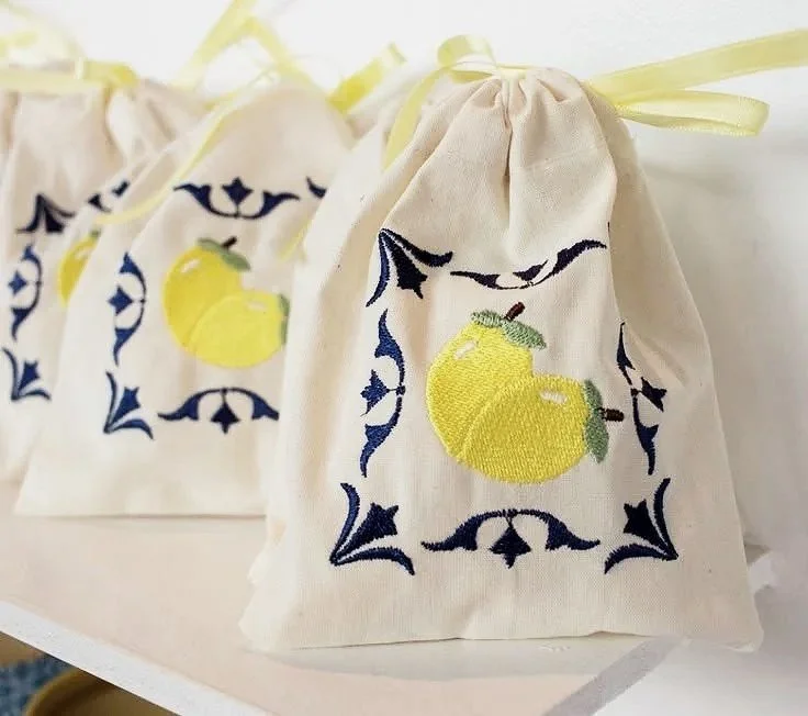 Cream-colored fabric gift bags with yellow apples and blue decorative border designs, tied with light yellow ribbons.