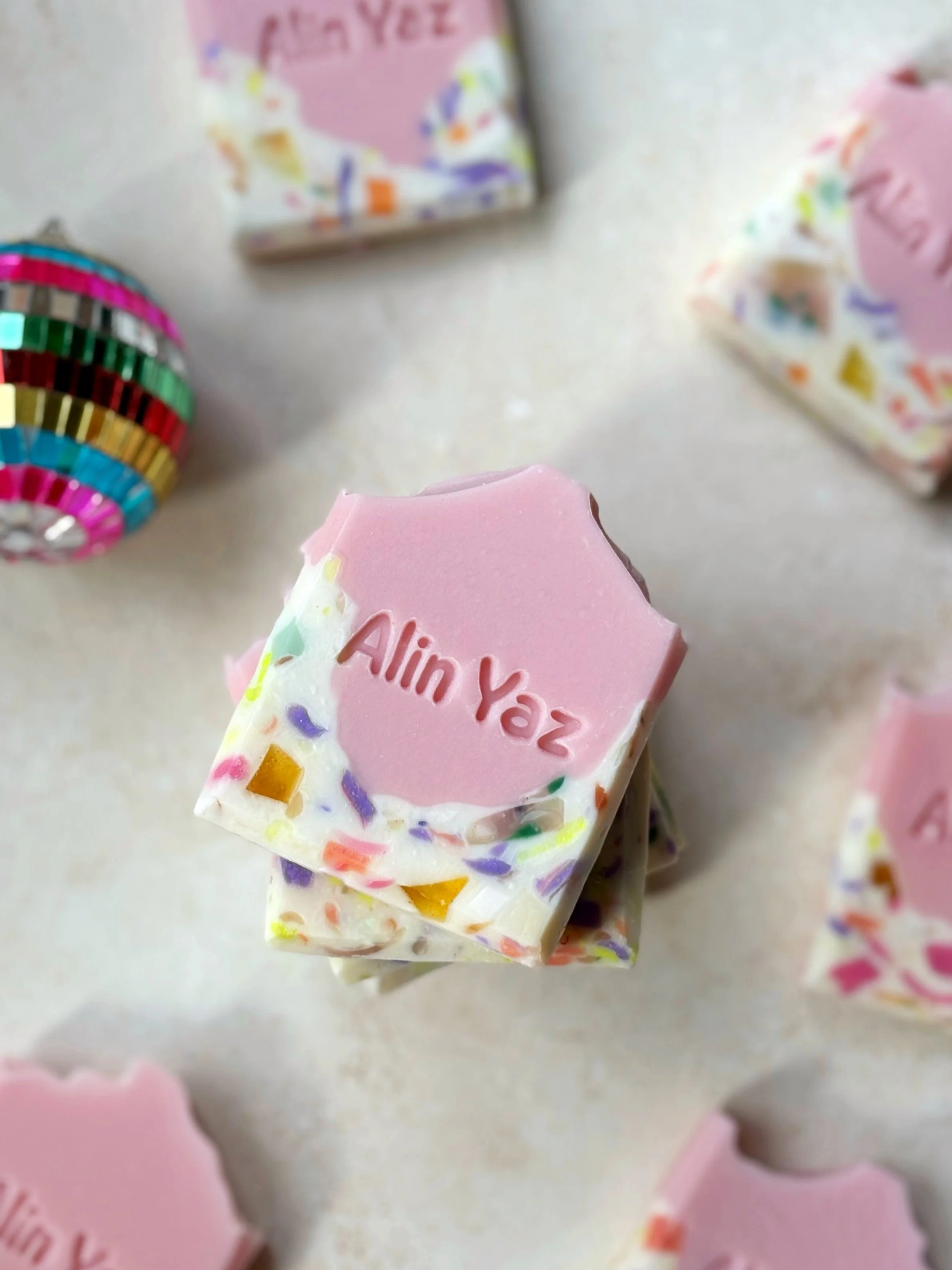 Square-shaped soap with a pink top layer engraved with the name 'Alin Vaz,' white base with colorful confetti-like soap pieces, positioned on a light countertop among other similar soaps and colorful decorative items.