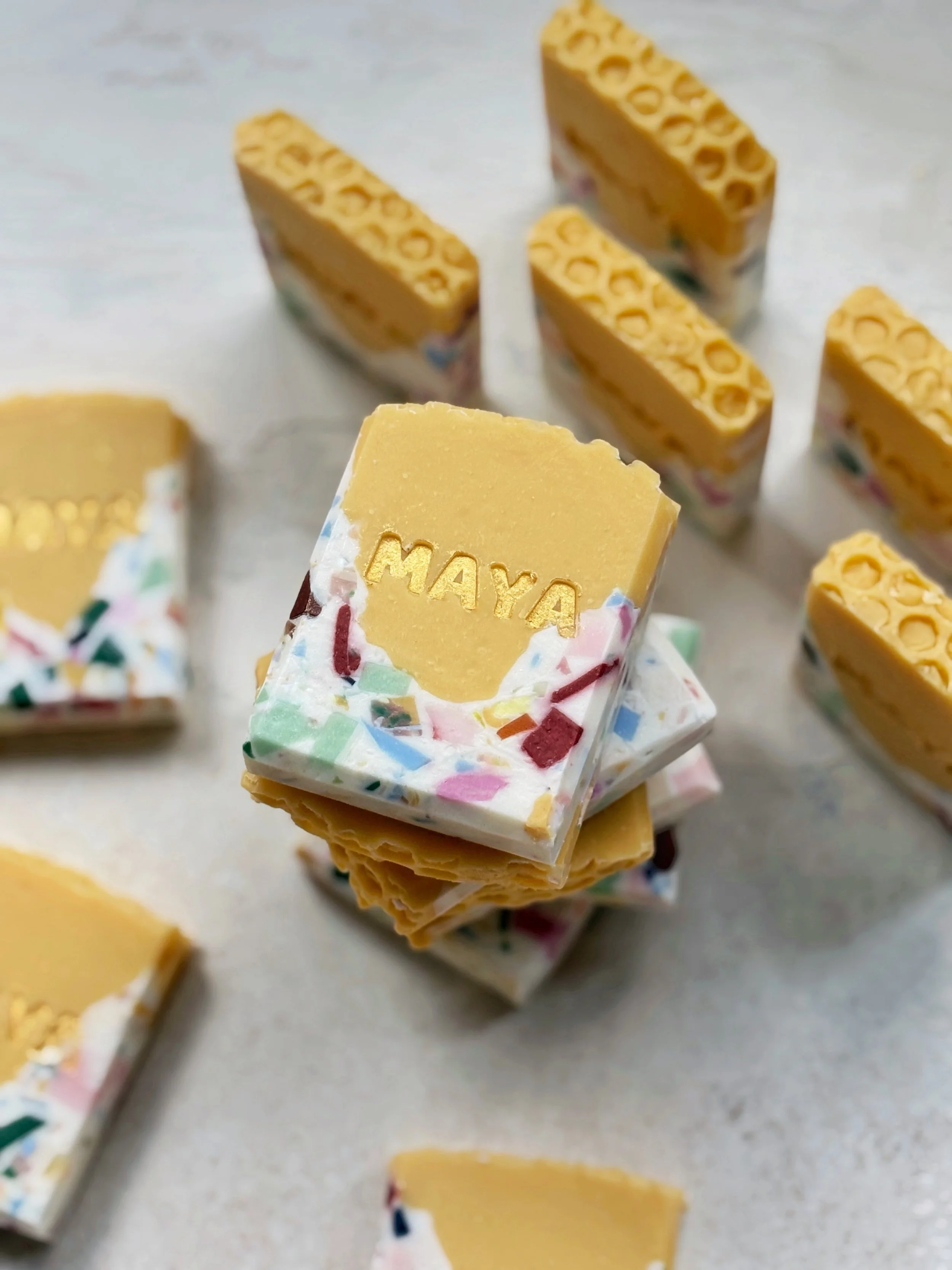 Stack of handmade soap bars with a yellow top layer and colorful sprinkle-like decoration around the edges, with one soap bar embossed with the name "MAYA."