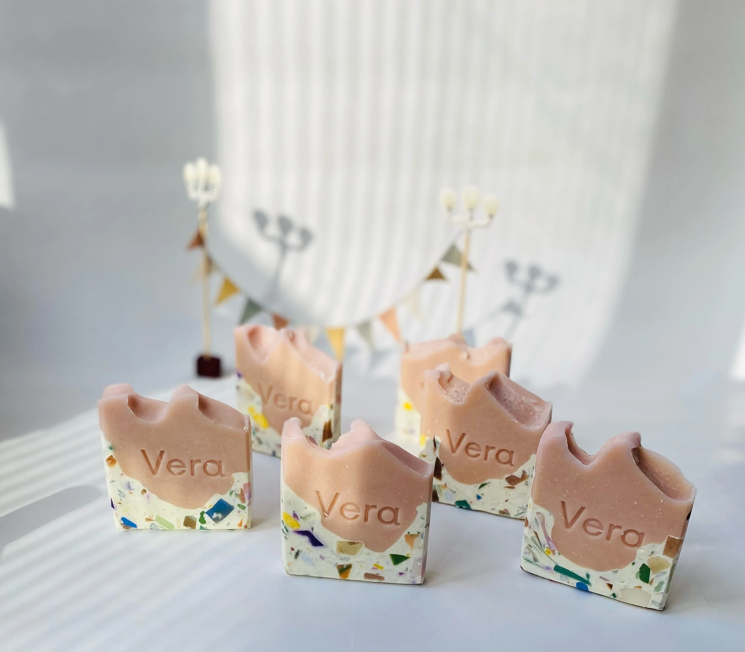 Six handmade soap bars with the name 'Vera' engraved on them, featuring a two-tone design with a pink top layer and a multicolored terrazzo-style bottom layer, arranged on a white surface with a blurred background of a window and decorative bunting.
