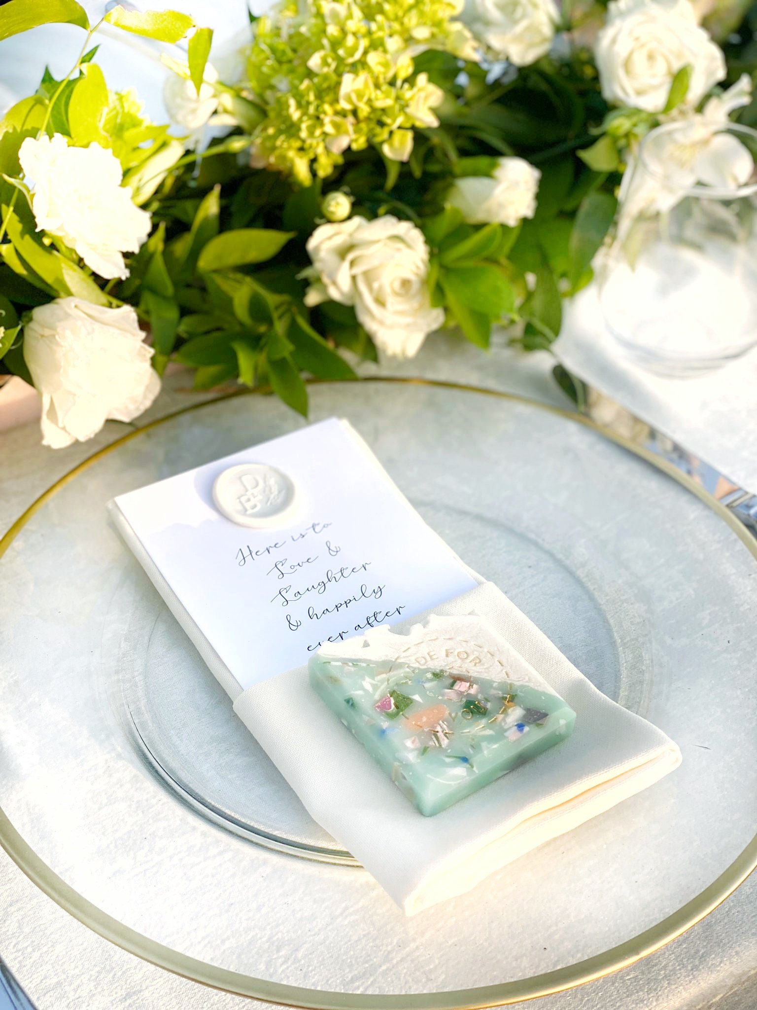 A wedding table setting featuring a bouquet of white flowers, a clear glass or crystal plate with a white napkin underneath, a handwritten note, and a soap with colorful stones inside, placed in front of the flowers.