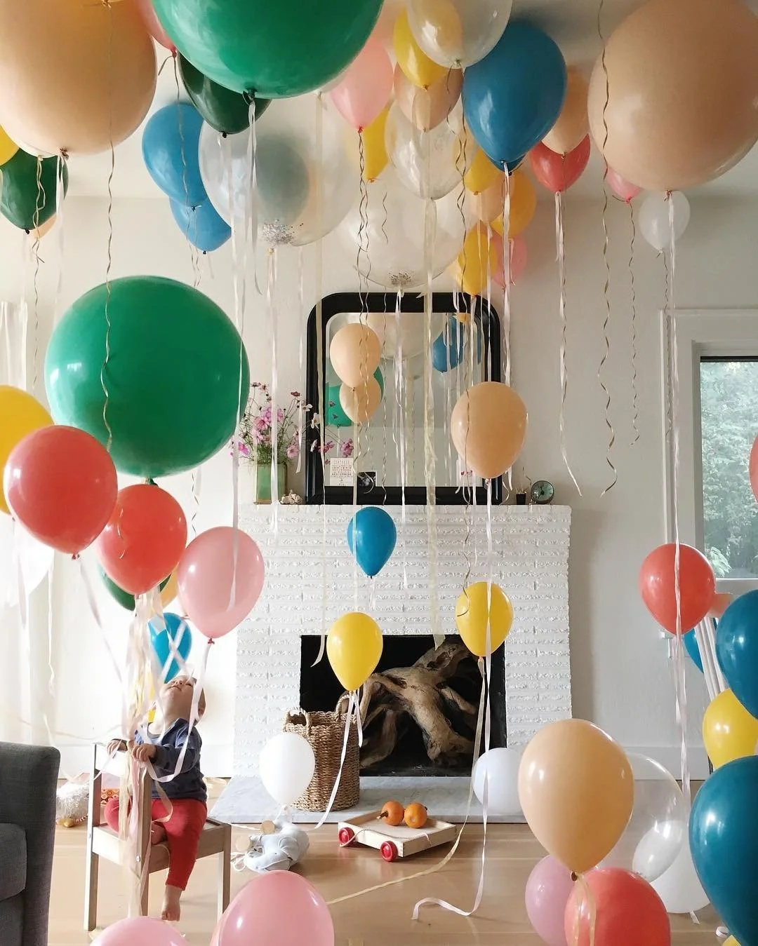 Room decorated with colorful balloons floating in the air, with a child reaching up among them. The room has a white brick fireplace with a mirror above it, a vase with pink flowers, and a window letting in natural light.