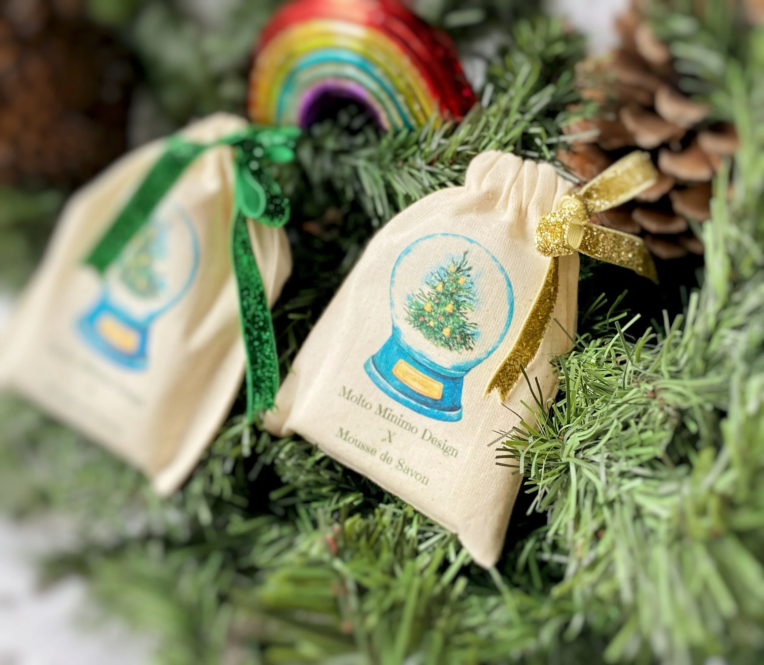 Decorative fabric pouches with Christmas tree prints, tied with green and gold ribbons, hanging on a Christmas tree branch with a rainbow ornament in the background.