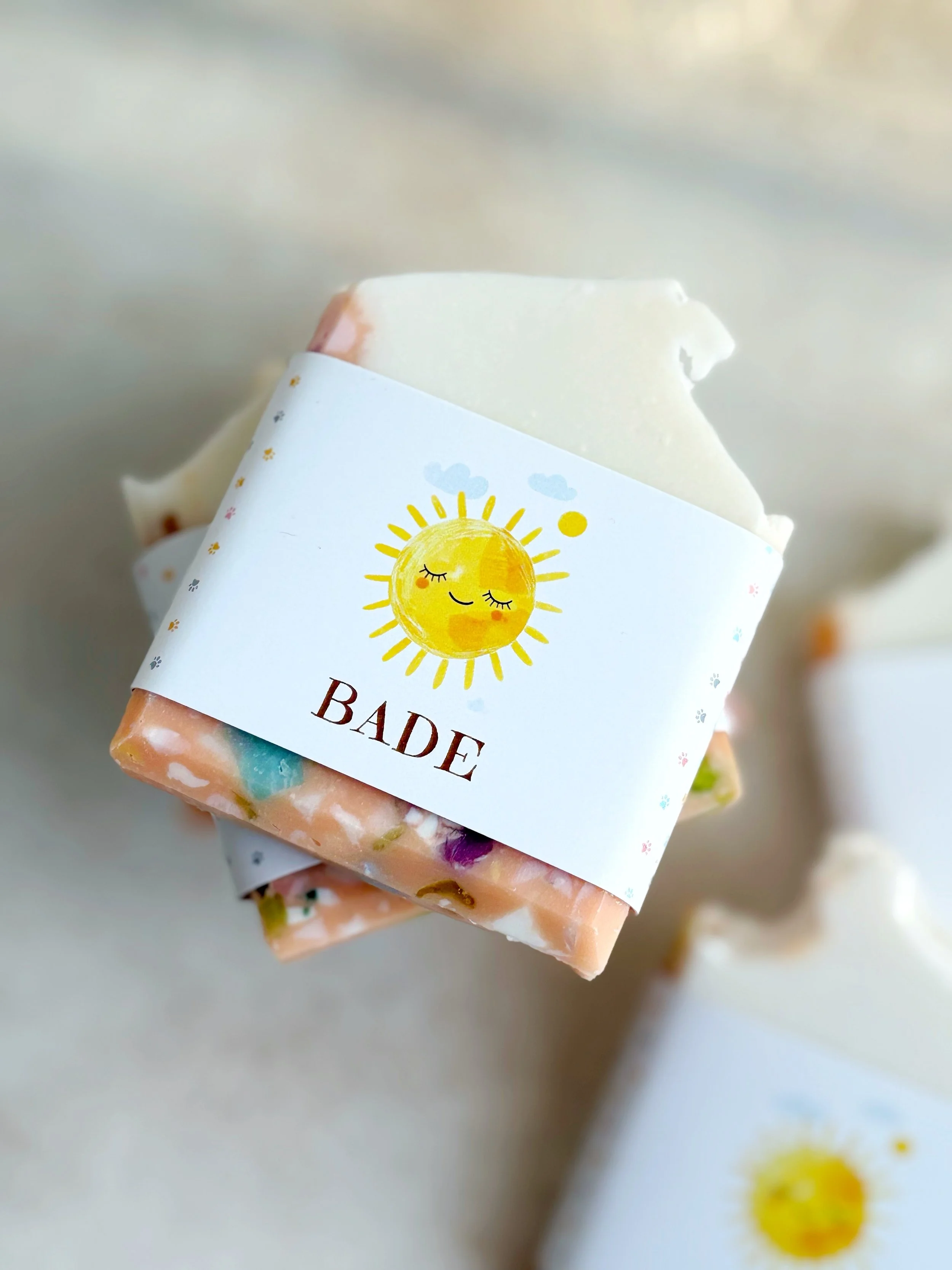 A bar of soap with a birthday cake scent, wrapped with a label featuring a smiling sun with closed eyes and the word "Bade" beneath it.