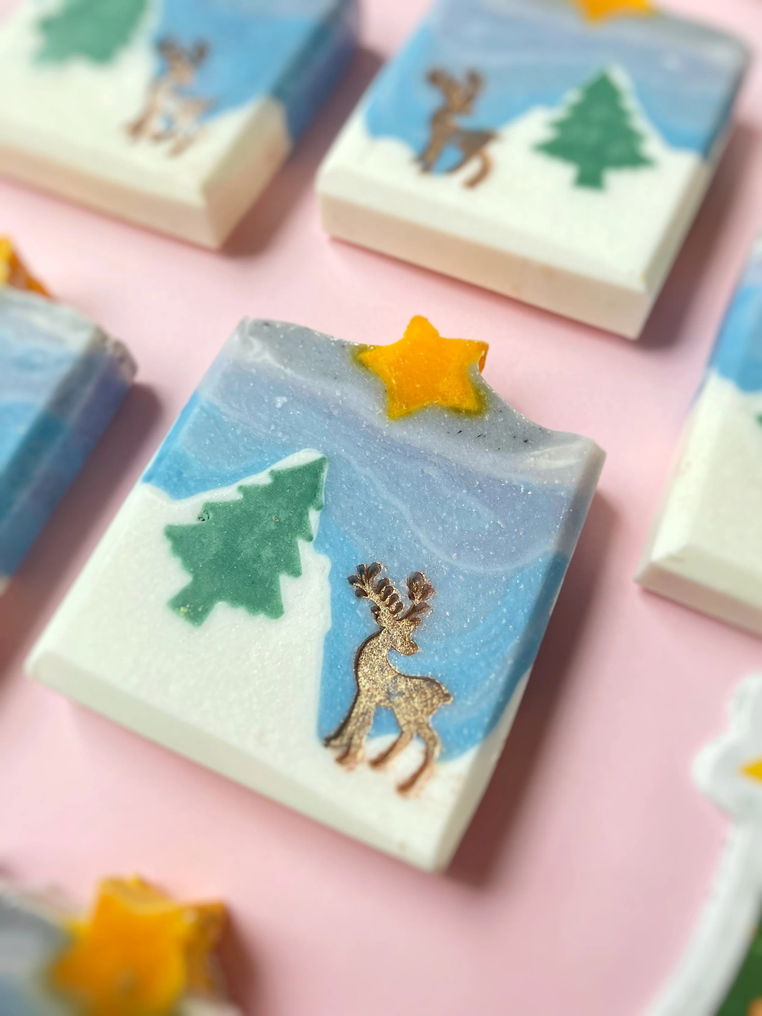 Decorative soap with a winter scene featuring a reindeer, pine trees, and a yellow star on top, set against a snowy landscape background.