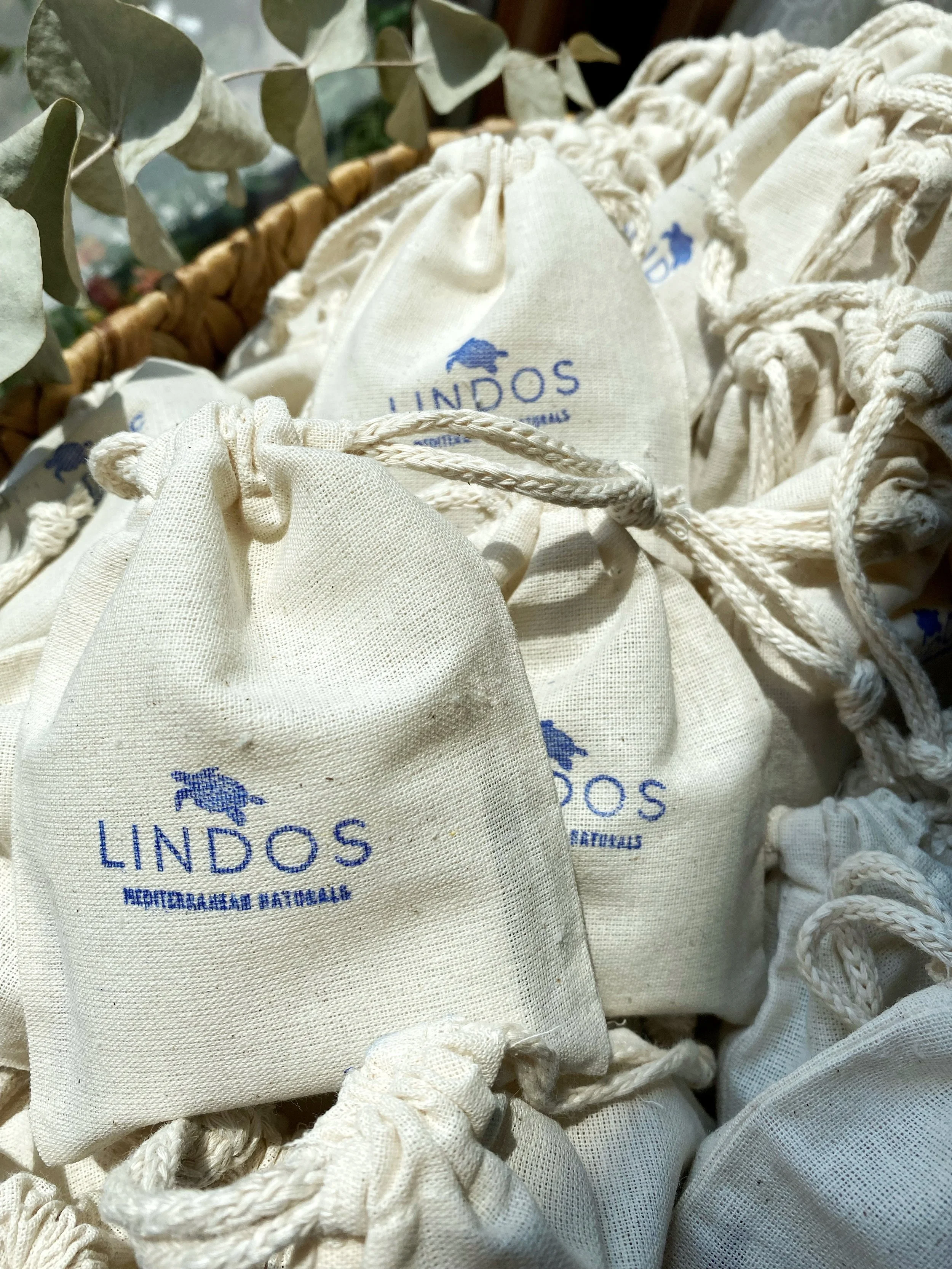 Multiple beige fabric drawstring bags with blue logo and text reading 'LINDOS' and 'AROMATICA NATURAL' placed in a wicker basket, with some green foliage visible in the background.