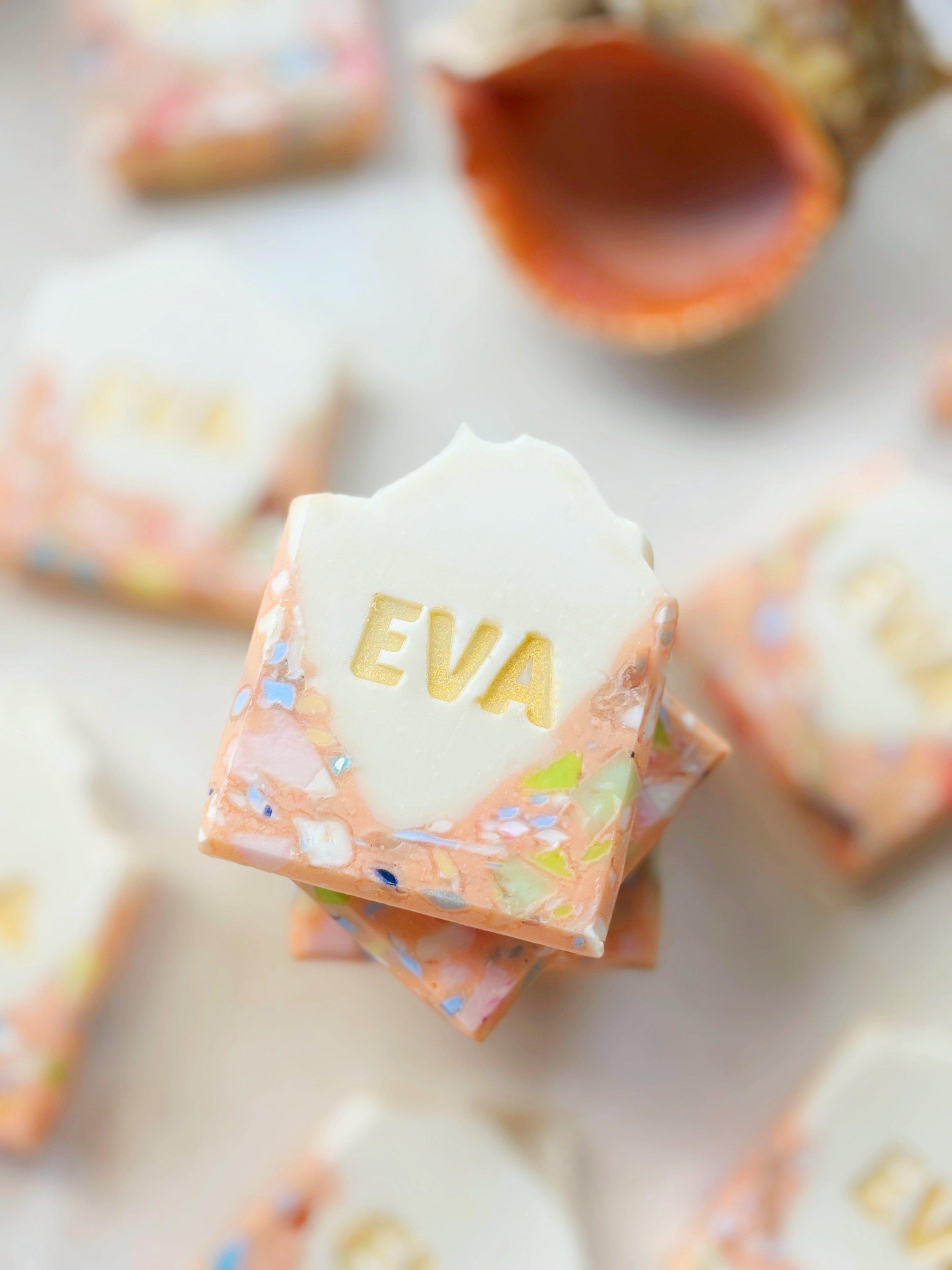 Personalized soap with the name 'EVA' engraved on top, surrounded by other colorful soaps, some with peeling or broken edges, on a light-colored surface.