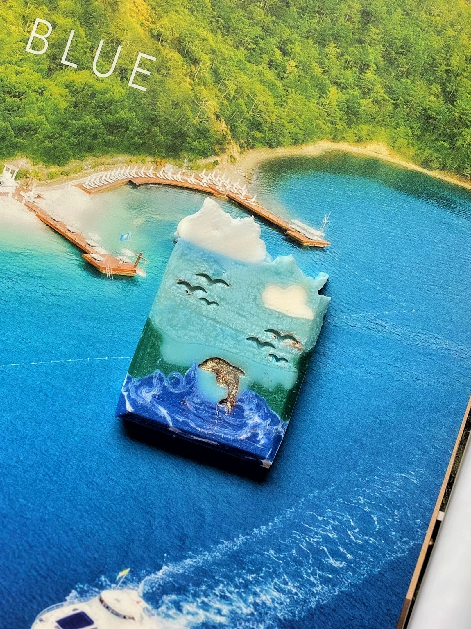 A colorful soap with ocean, clouds, and bird designs overlays a photo of a lake or ocean with a dock and lounge chairs on the shoreline.