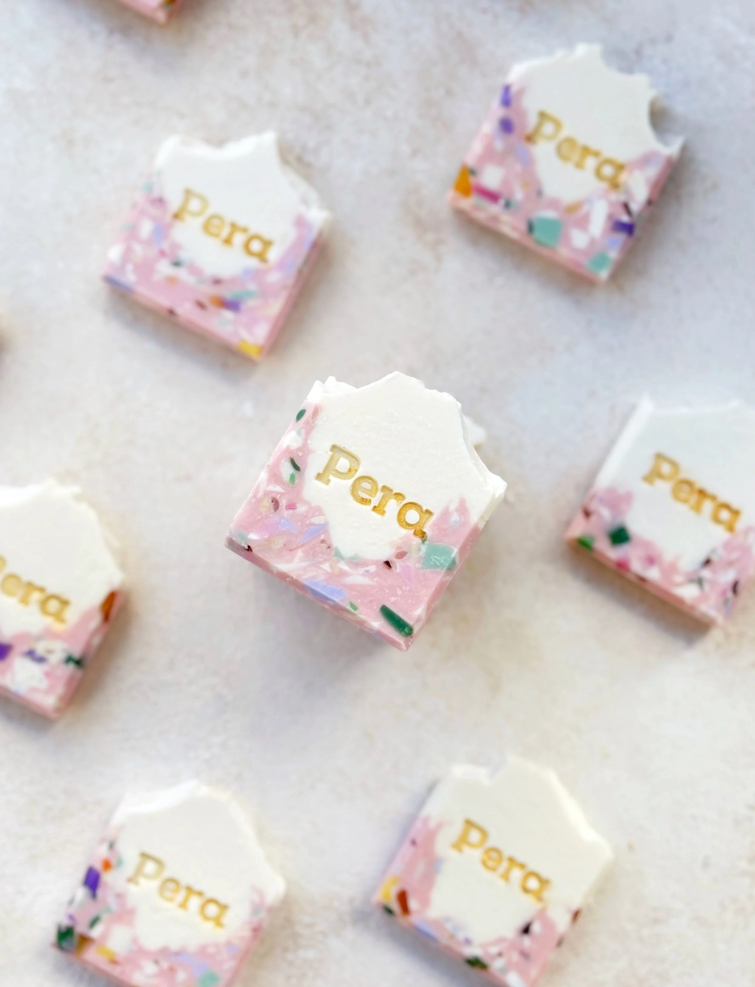 Pink and white terrazzo-style soap bars with the brand name 'Pera' embossed in gold, arranged on a white surface.