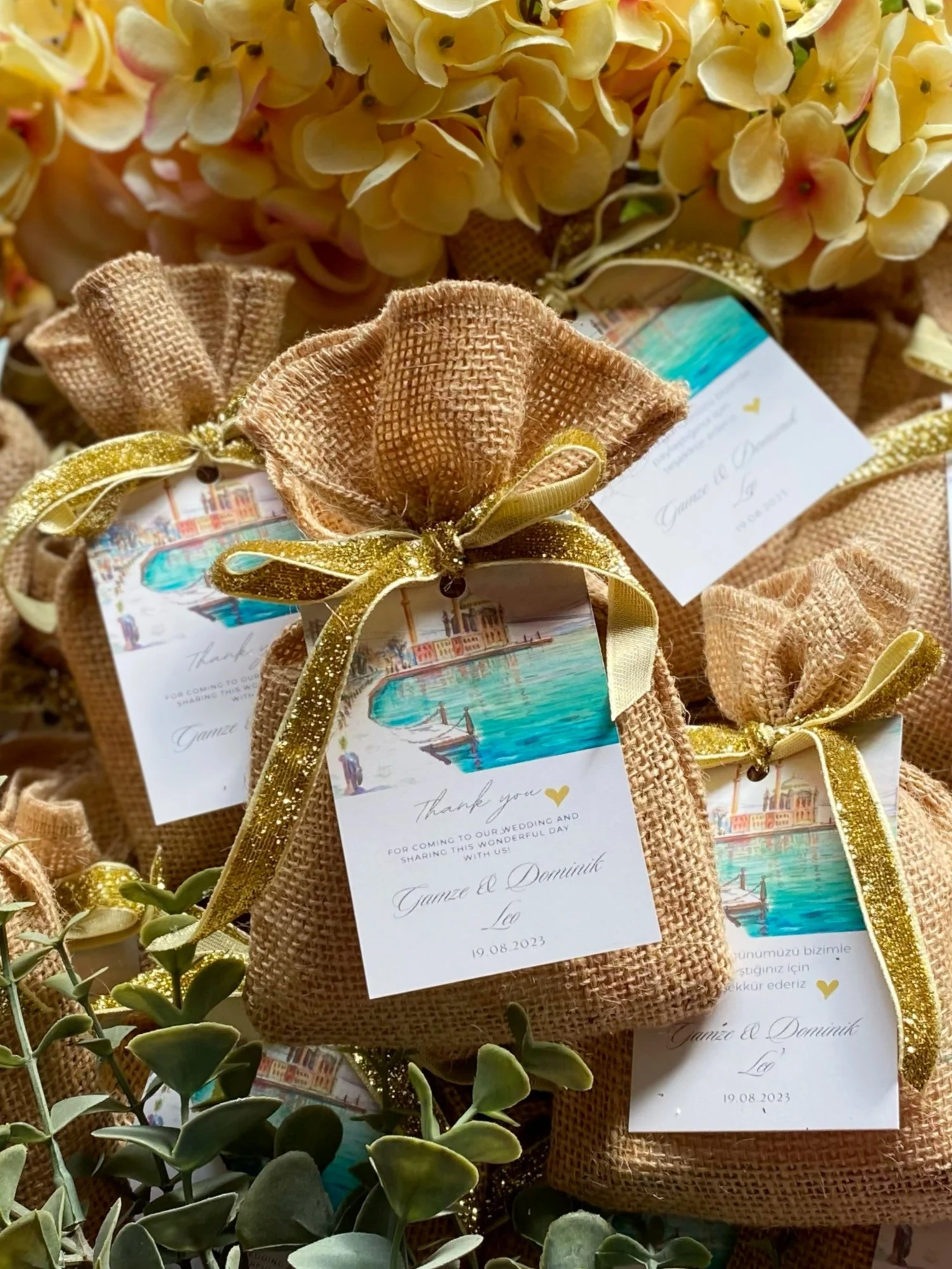 Small burlap pouches with gold ribbon and thank you cards, surrounded by yellow flowers and green foliage.