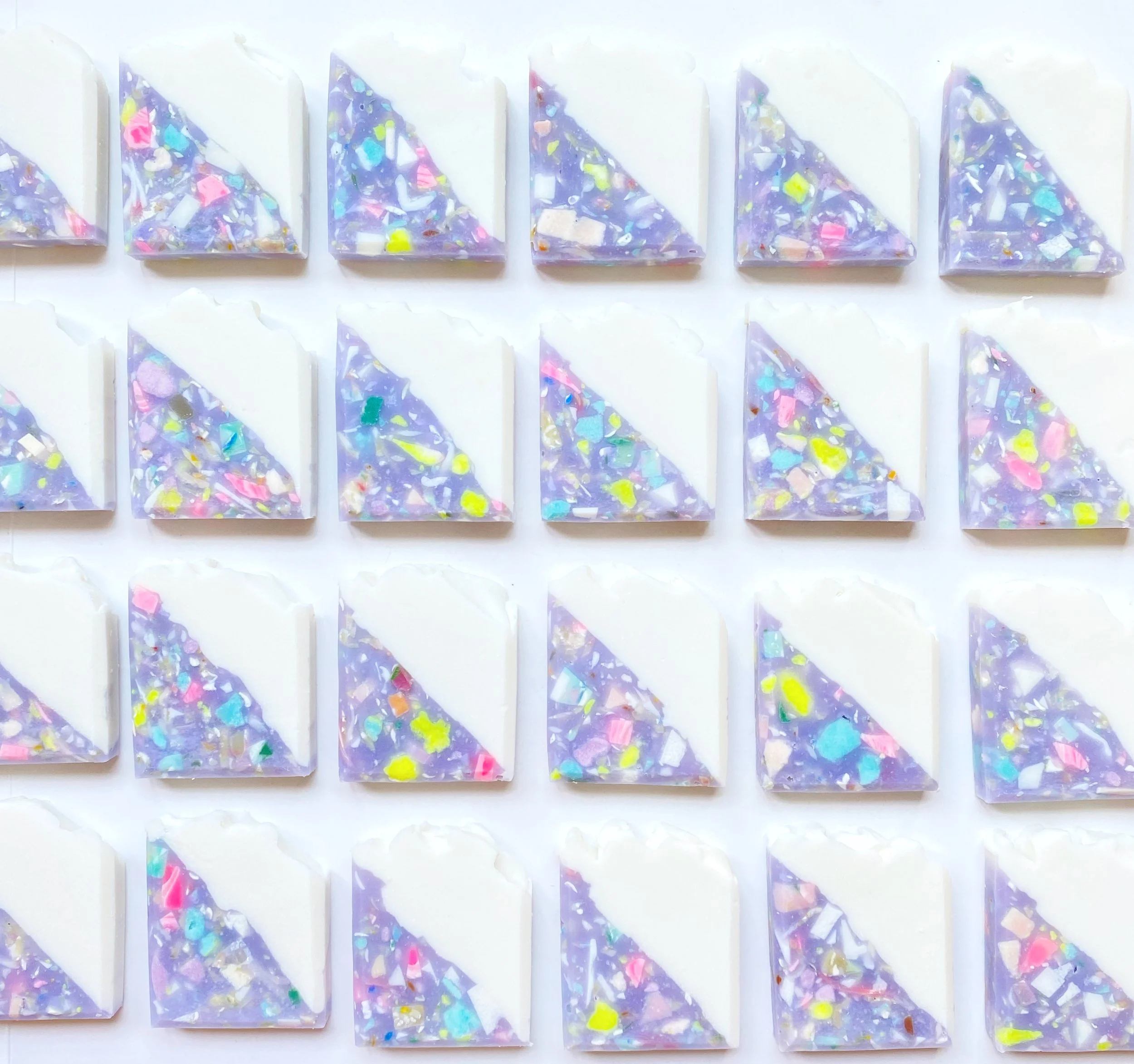 Assorted square and triangular pieces of soap with iridescent, colorful, glittery designs arranged in rows on a white surface.