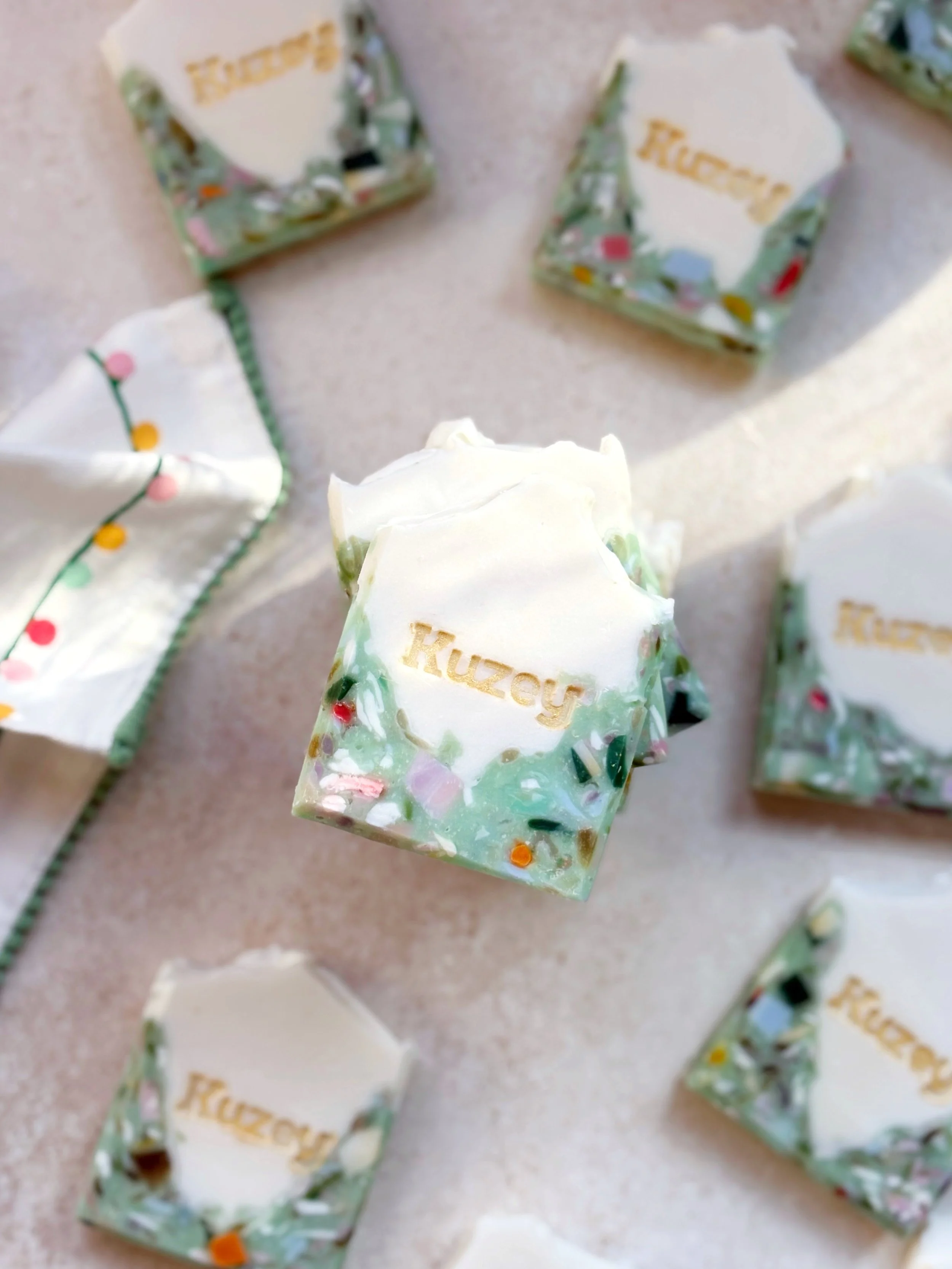 A bar of handmade soap labeled 'Ruzzy' with a white top and a colorful, glittery base, surrounded by similar soap and a cloth with Christmas lights design on a neutral surface.