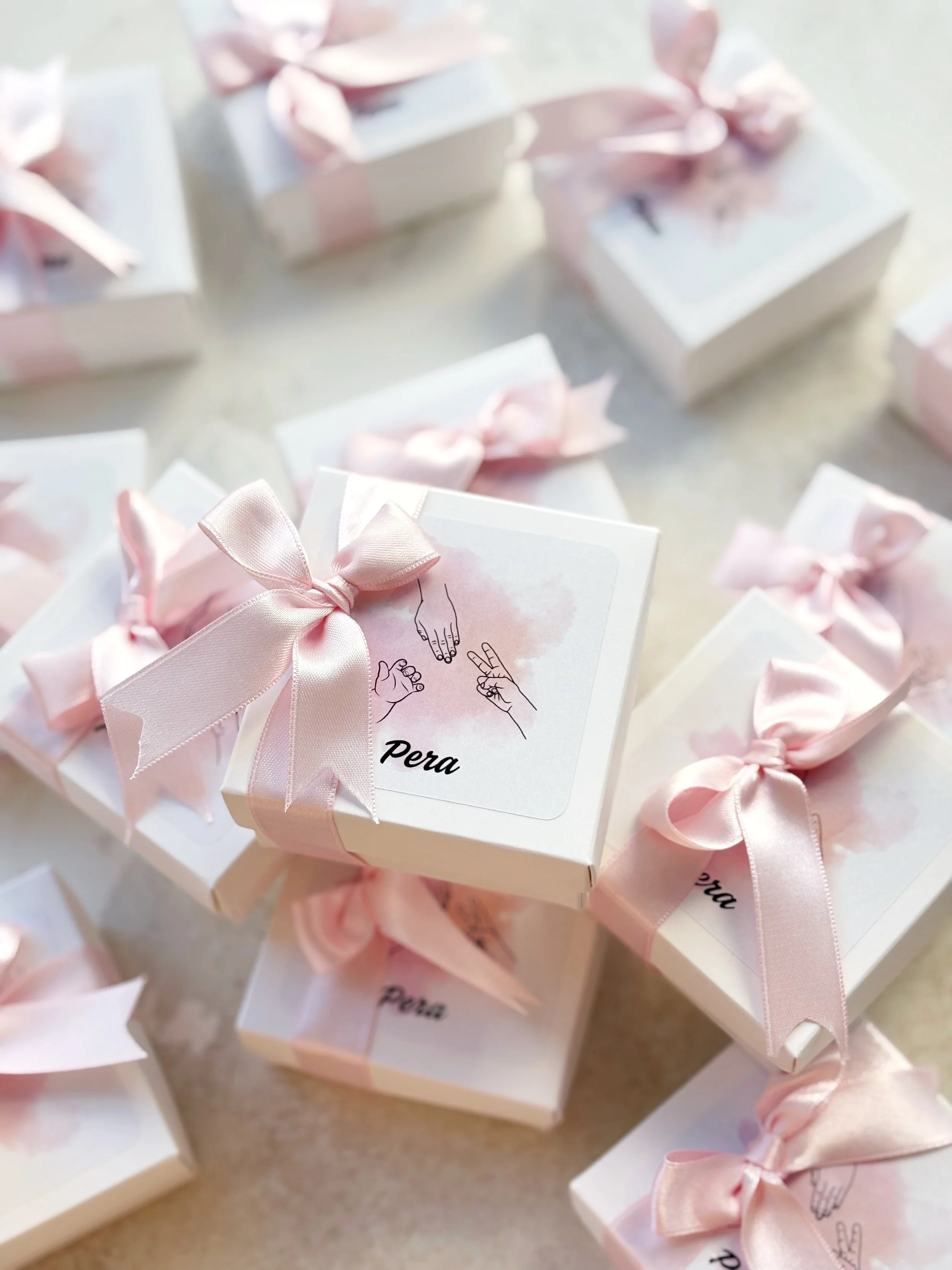 Multiple white gift boxes decorated with pink satin ribbons and bows, with one box featuring an illustration of hands making different sign language gestures and the word 'Pera' on the label.