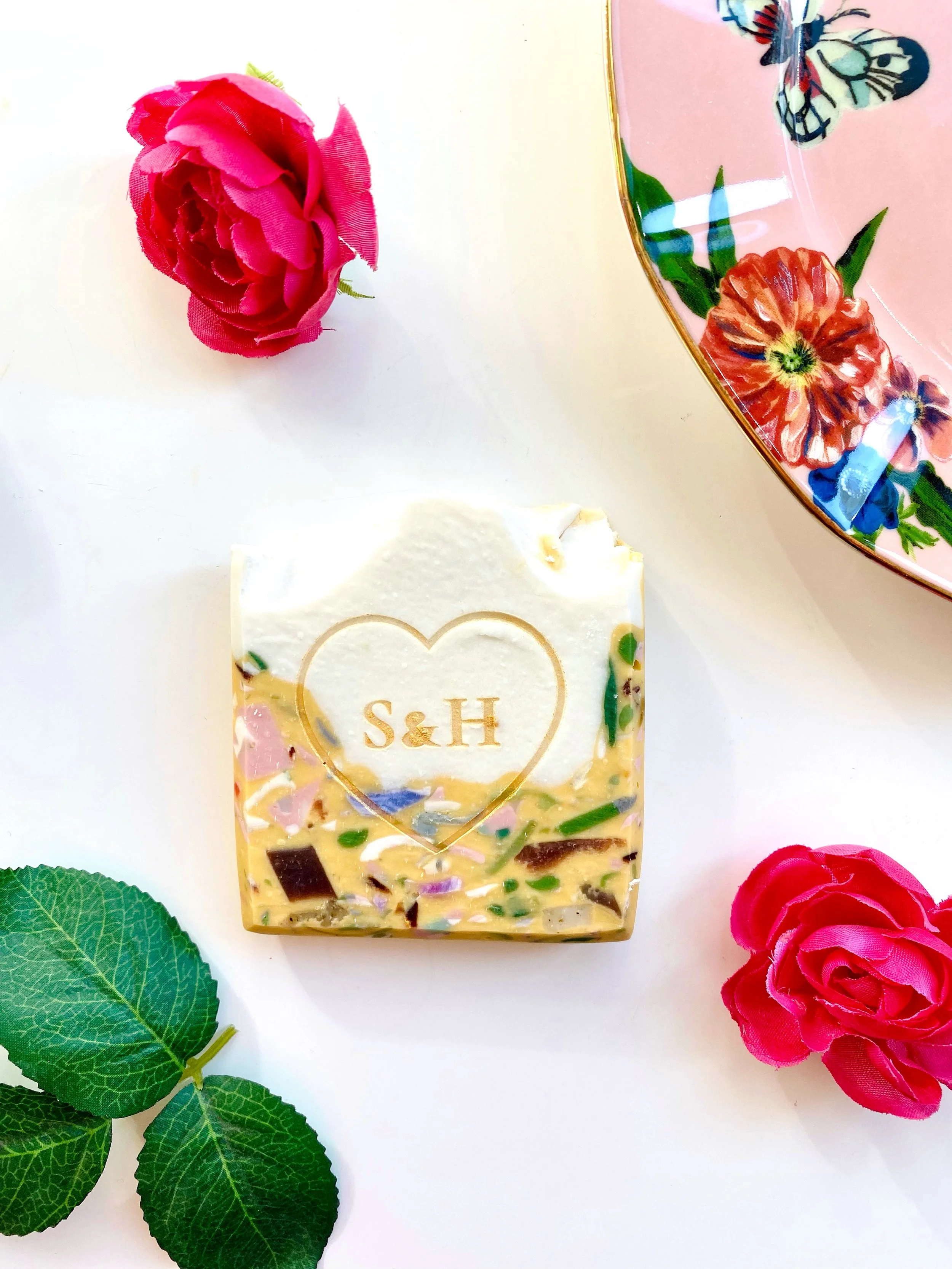 Decorative soap with gold lettering 'S & H' inside a heart shape, pink roses, green leaves, and floral-patterned plate on a white surface.