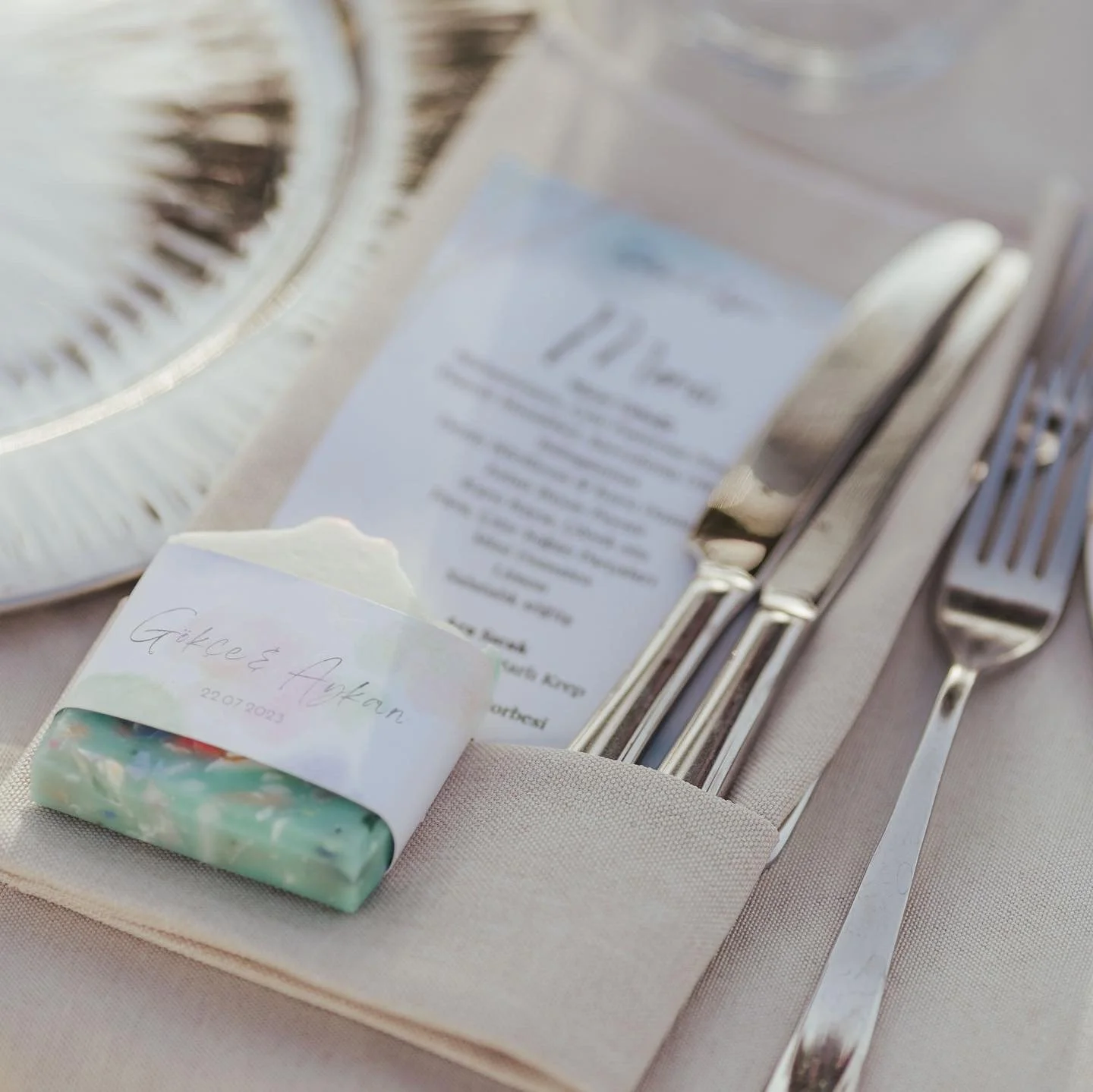Set table with utensils, a menu, a soap bar with a handwritten name tag, and a decorative plate in the background.