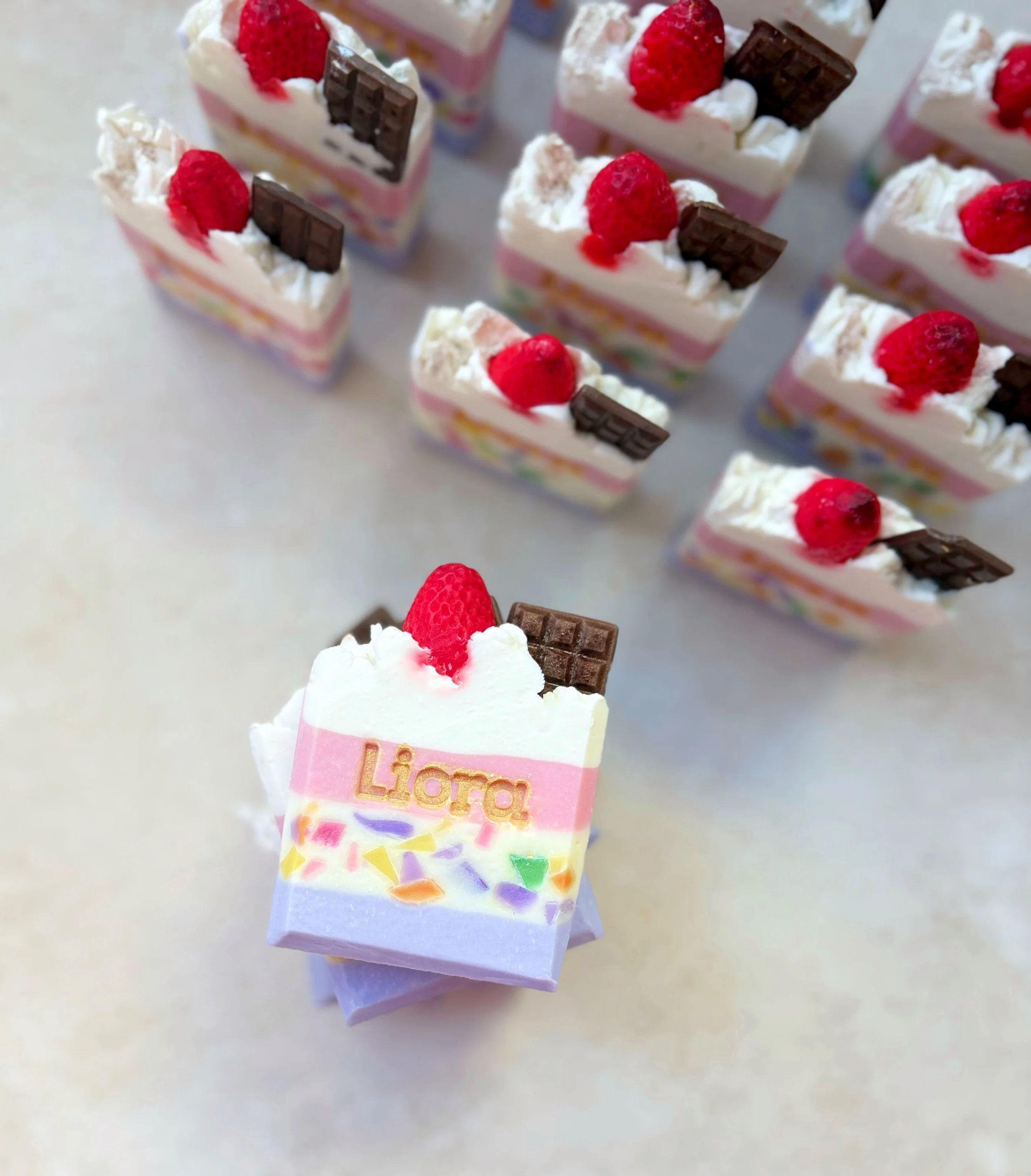 Colorful layered soap bars topped with white cream, a raspberry, and a piece of chocolate, with the front soap bar bearing the word "Liora".