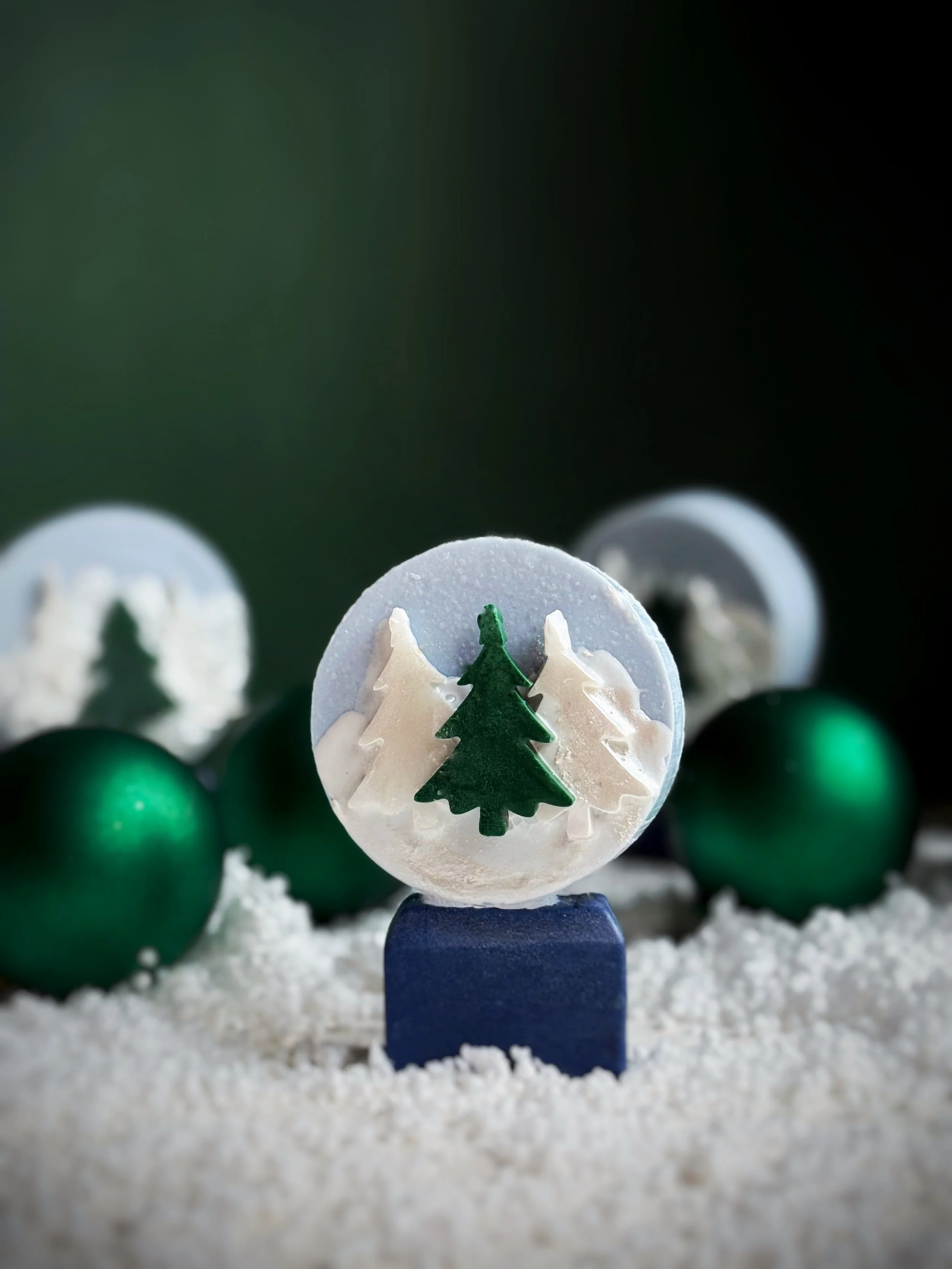 Close-up of Christmas-themed candy with green tree design, surrounded by green ornaments on white faux snow, with a dark background.