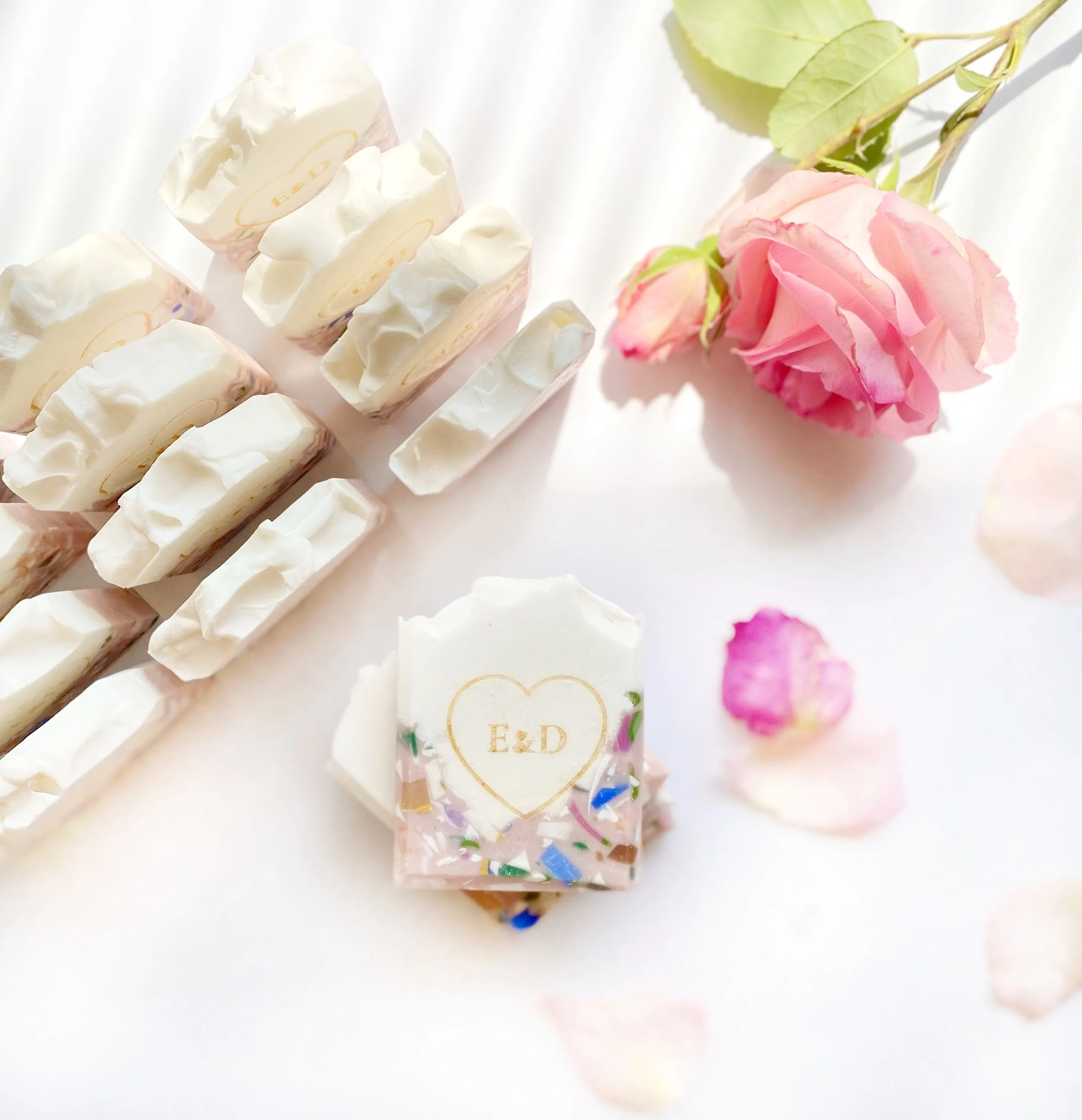 A close-up of white rectangular chocolates with ornate detailing, a pink rose with green leaves, and pink rose petals scattered around on a white surface.