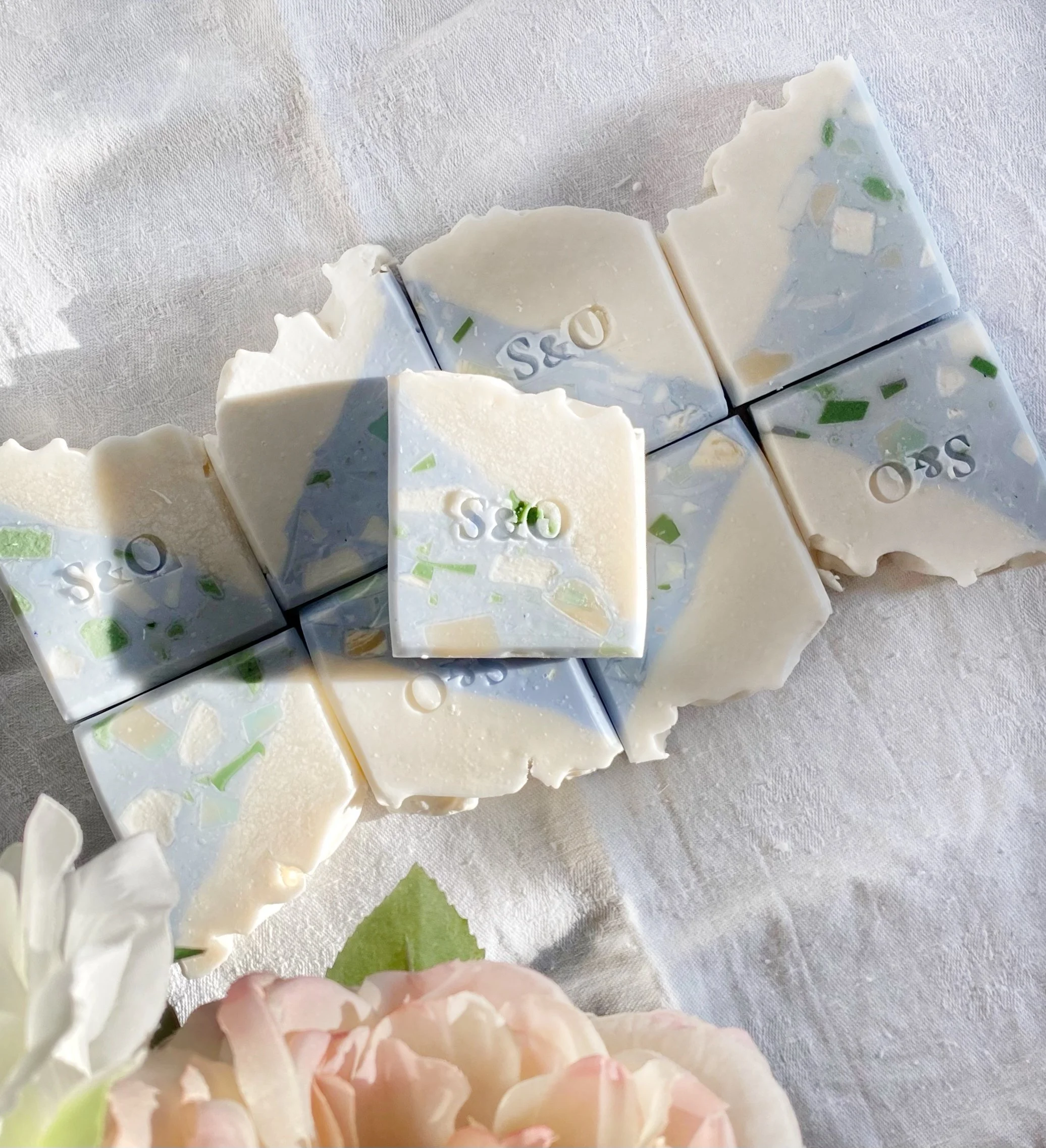 Pieces of white soap with green and yellow flakes and embossed design, on white fabric background, with pink flowers in foreground.
