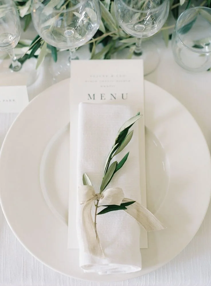 Elegant table setting with a white plate, cloth napkin decorated with a green leaf and ribbon, elegant menu card, and multiple wine glasses above.