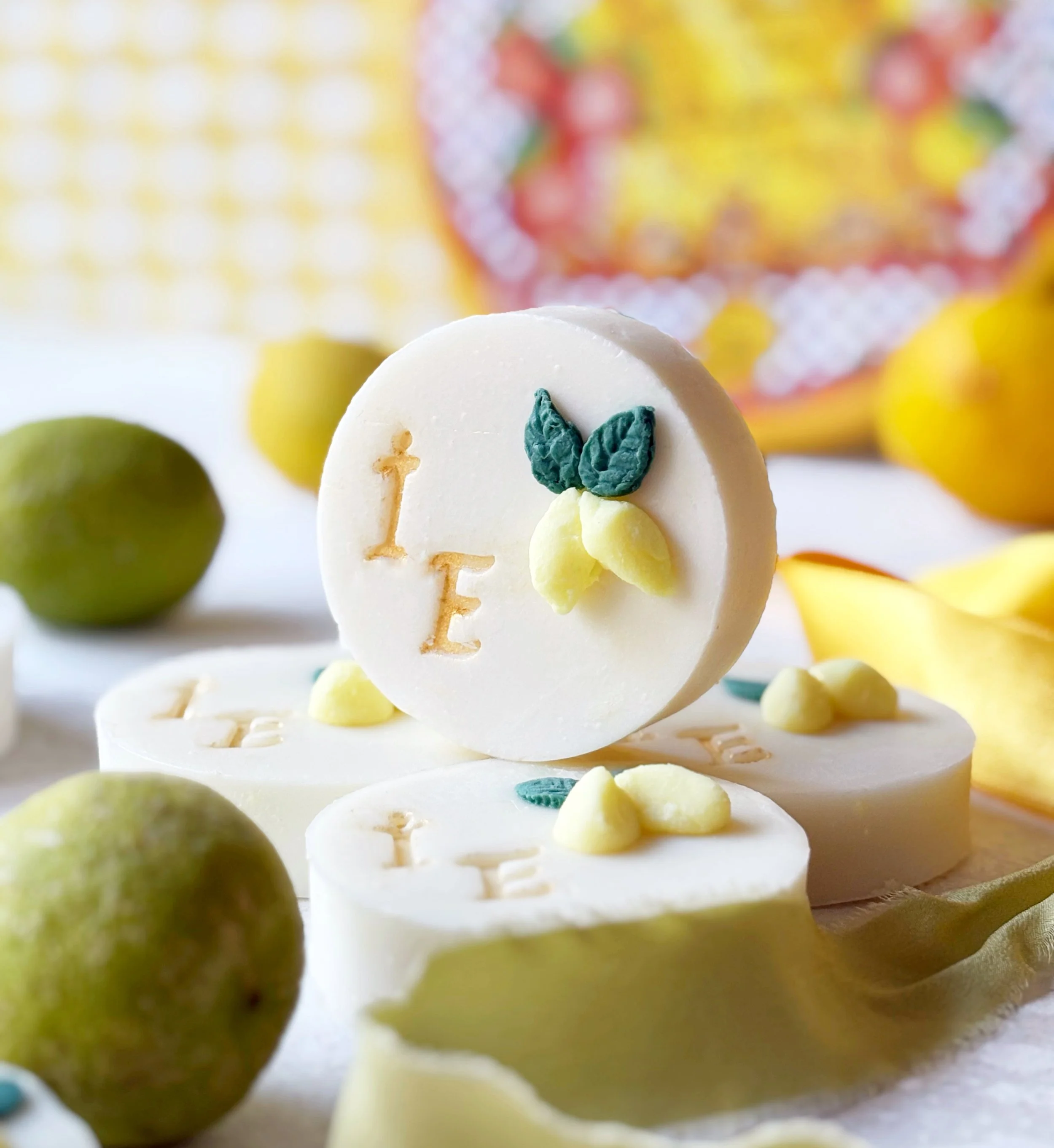 White circular soap with the word "LOVE" engraved and a lemon and green leaf decoration, surrounded by yellow lemons, a yellow flower, and blurred background.