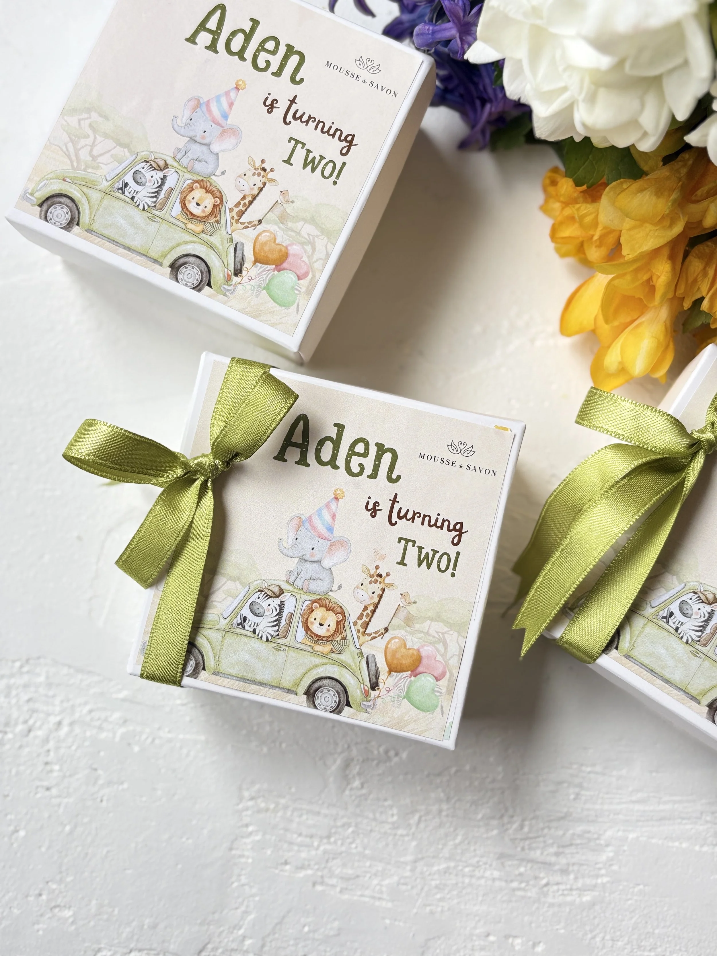 A gift box with a sticker that says "Aden is turning TWO!" featuring a cartoon illustration of a safari scene with a zebra, lion, elephant, and giraffe in a green car, decorated with balloons and a party hat.