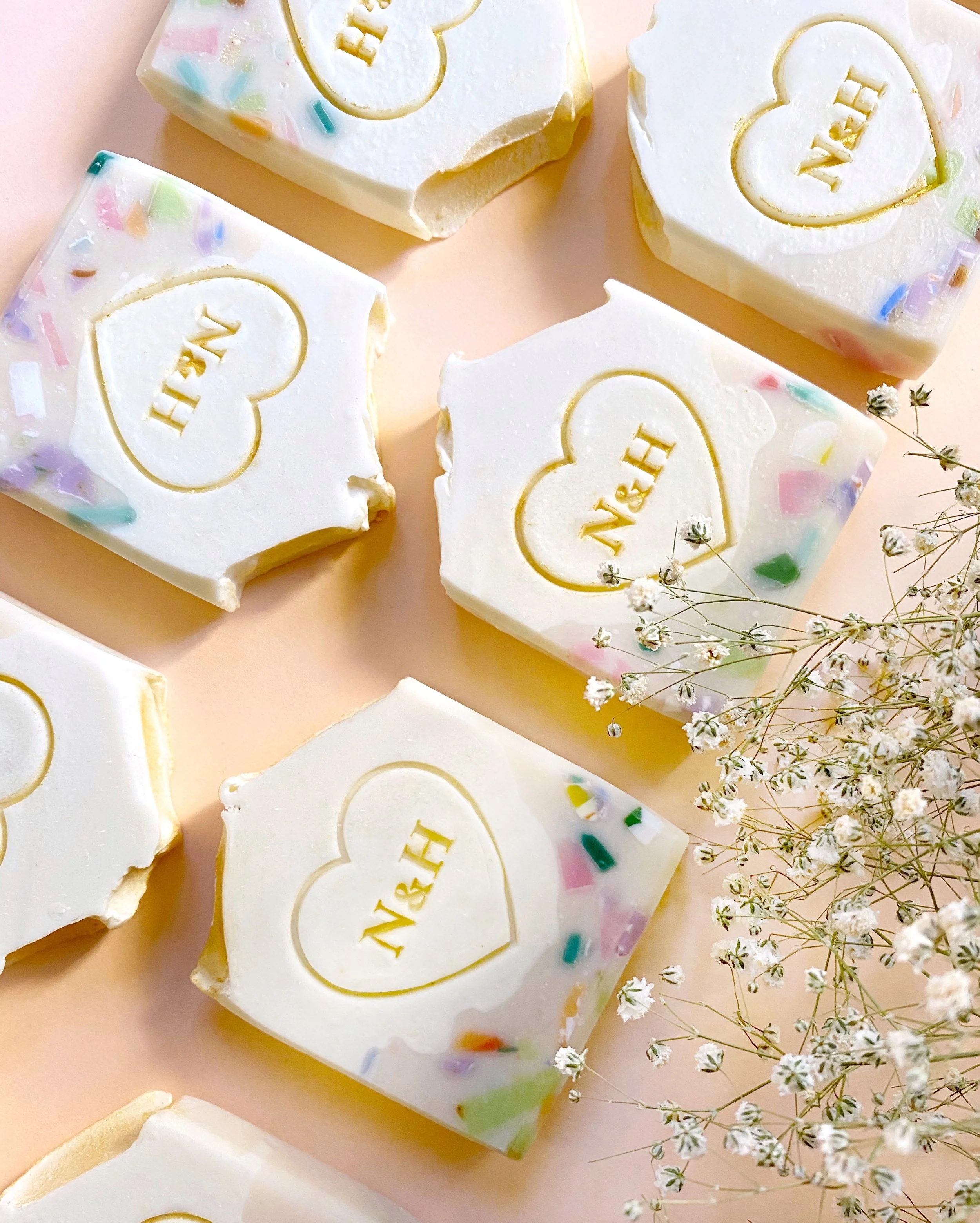 Bar of white soap with a heart-shaped embossed logo reading 'N&H', surrounded by small white flowers, on a pink background.