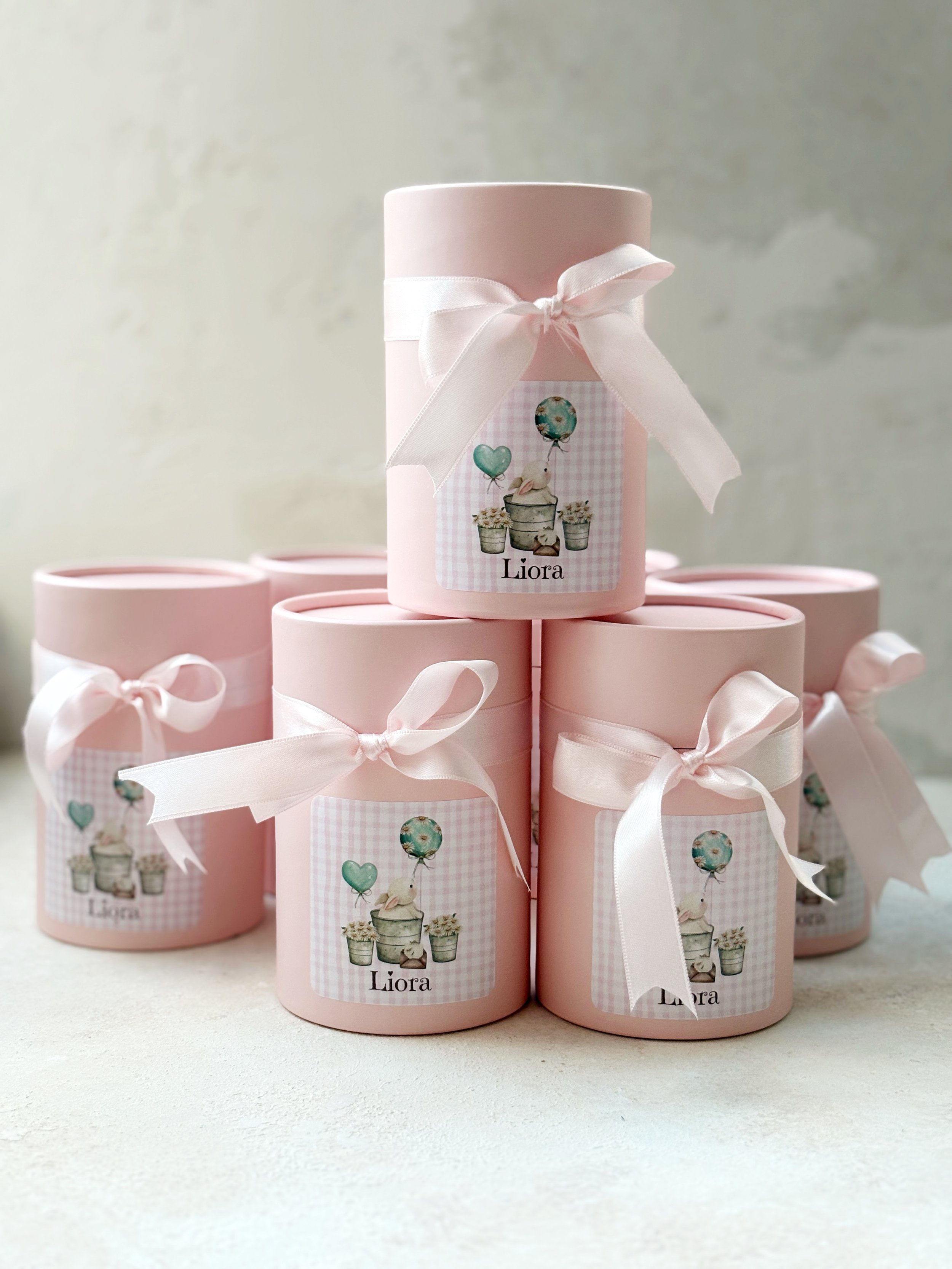 Pink cylindrical gift boxes with white ribbons and custom labels featuring a rabbit with balloons and potted plants, with the name 'Liora' on each label.