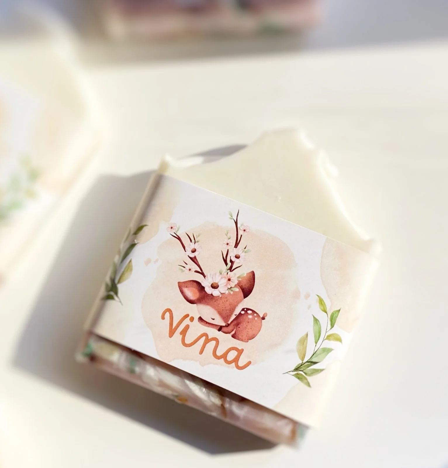 Small white box with a decorative label featuring a cartoon deer with antlers made of flowers, and the name 'Vina' written in orange cursive. The box is on a white surface with soft lighting.
