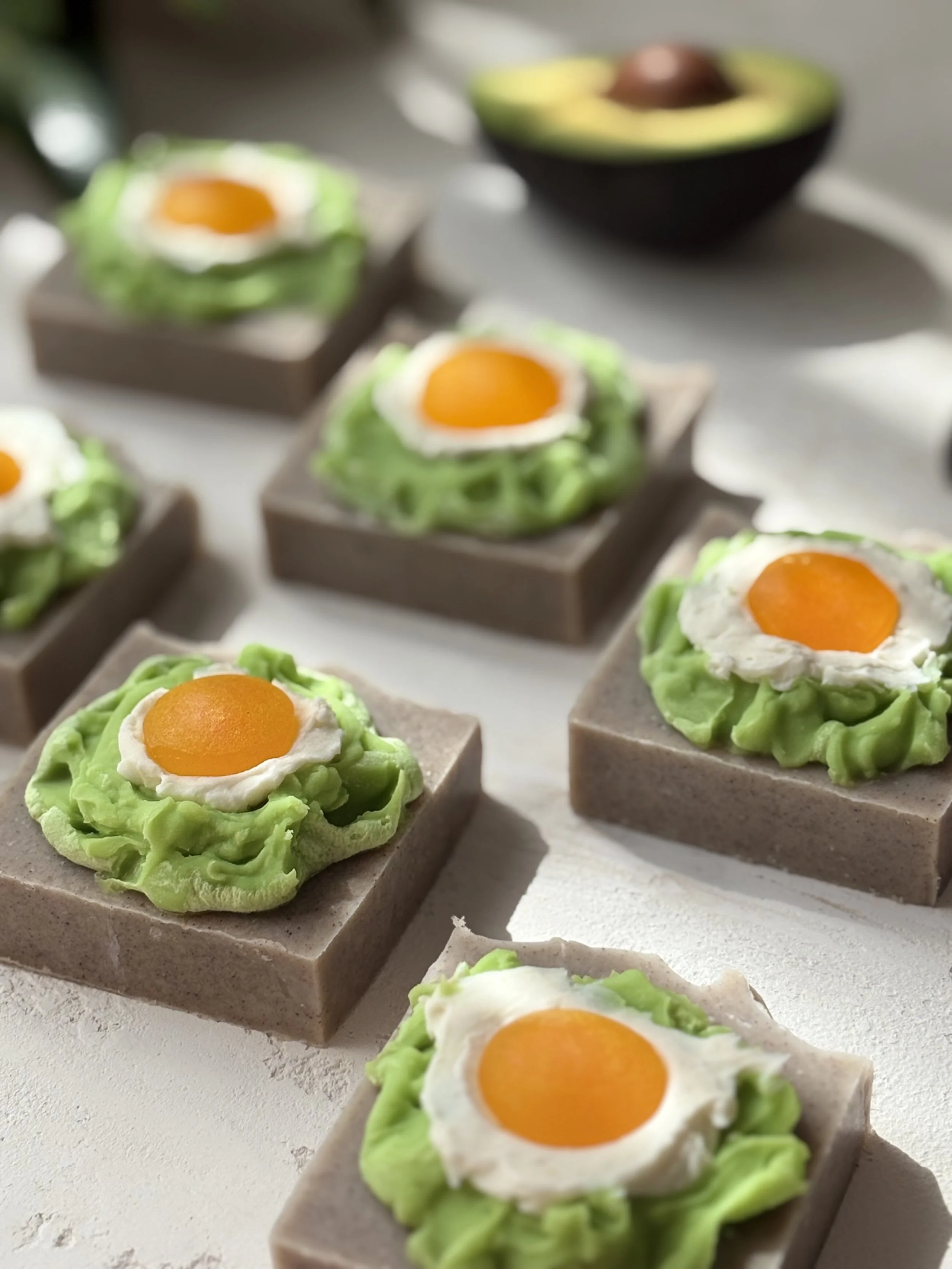 Several square-shaped soap bars topped with frosting and a small dollop of mashed avocado, decorated with a dollop of white cream and a small orange fruit slice on top, arranged on a white surface.