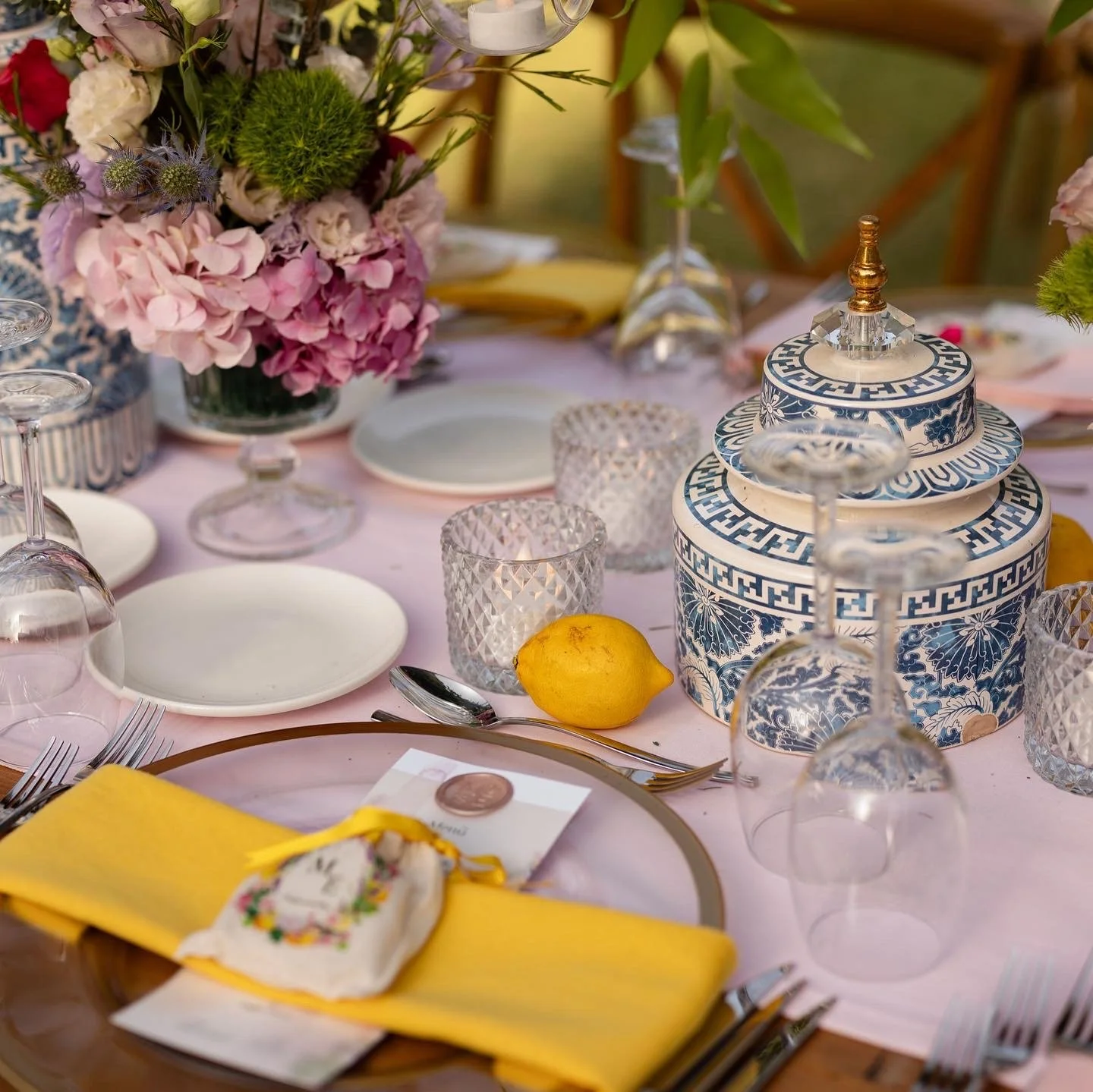 A tablescape with pink, purple, and green floral arrangements, lit candles in glass holders, white plates, gold-edged chargers, yellow napkins, clear glassware, a lemon, and a patterned blue and white decorative container.