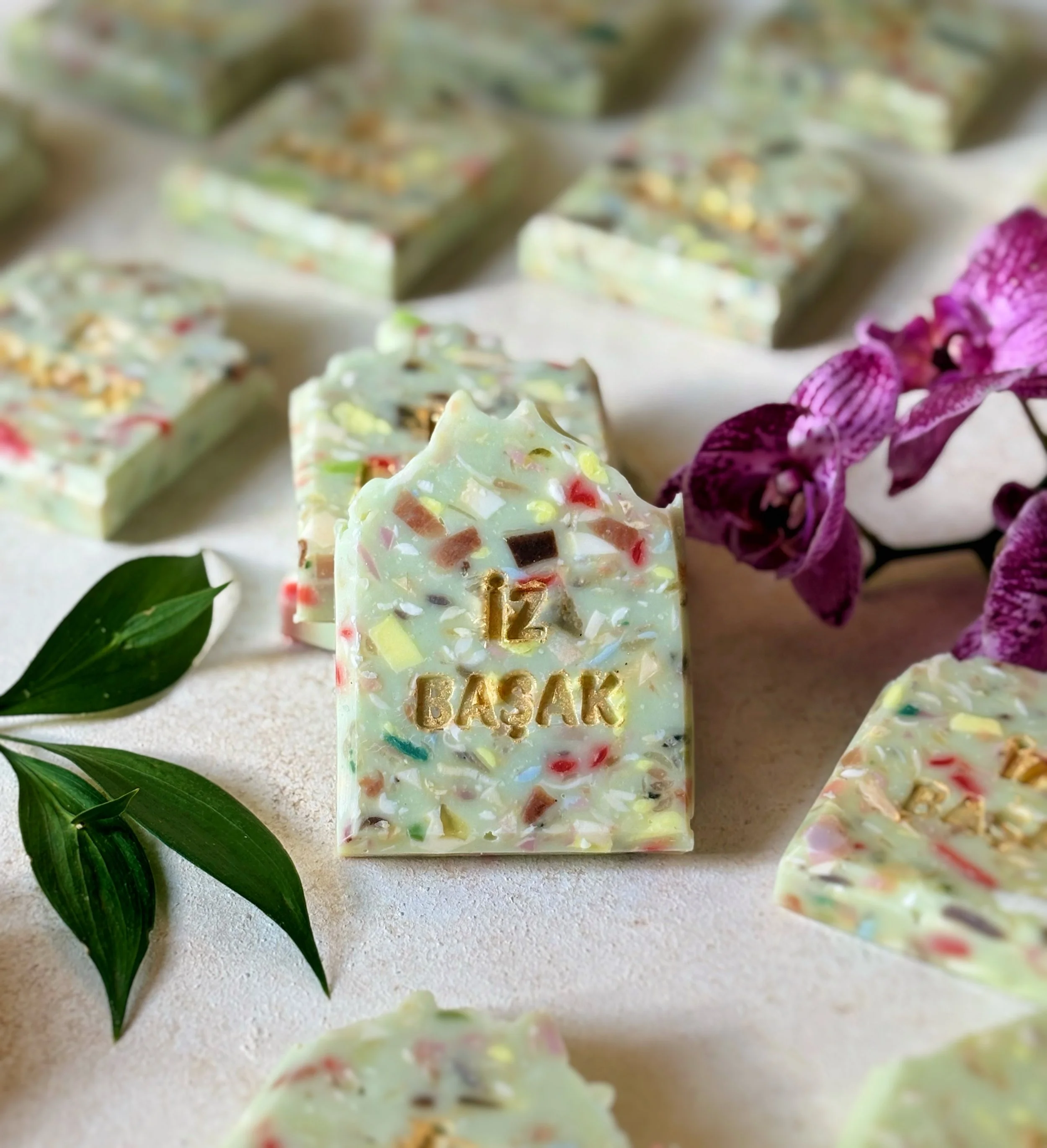 White chocolate with colorful candy pieces and gold lettering that reads "12 BASK" surrounded by purple orchids and green leaves.