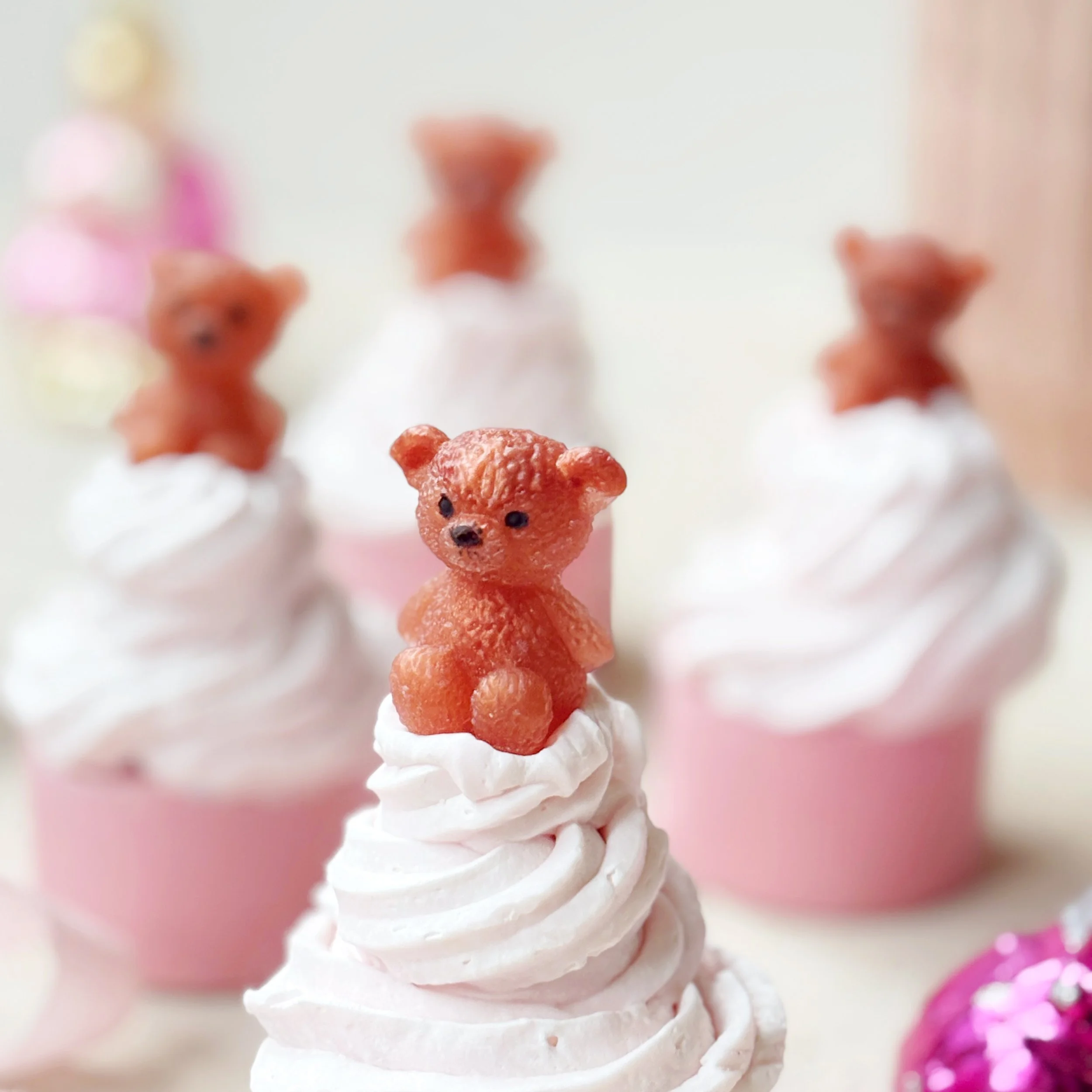 Pink cupcake soap liners with swirled white frosting topped with a small edible bear figurine, with three more similar cupcakes blurred in the background.