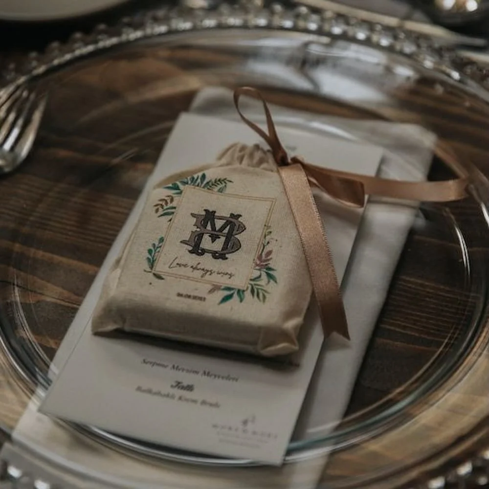 A gift wrapped in beige paper with a brown ribbon, placed inside a silver tray along with a printed menu or program. The gift has a fabric tag with an embroidered monogram surrounded by a floral design.