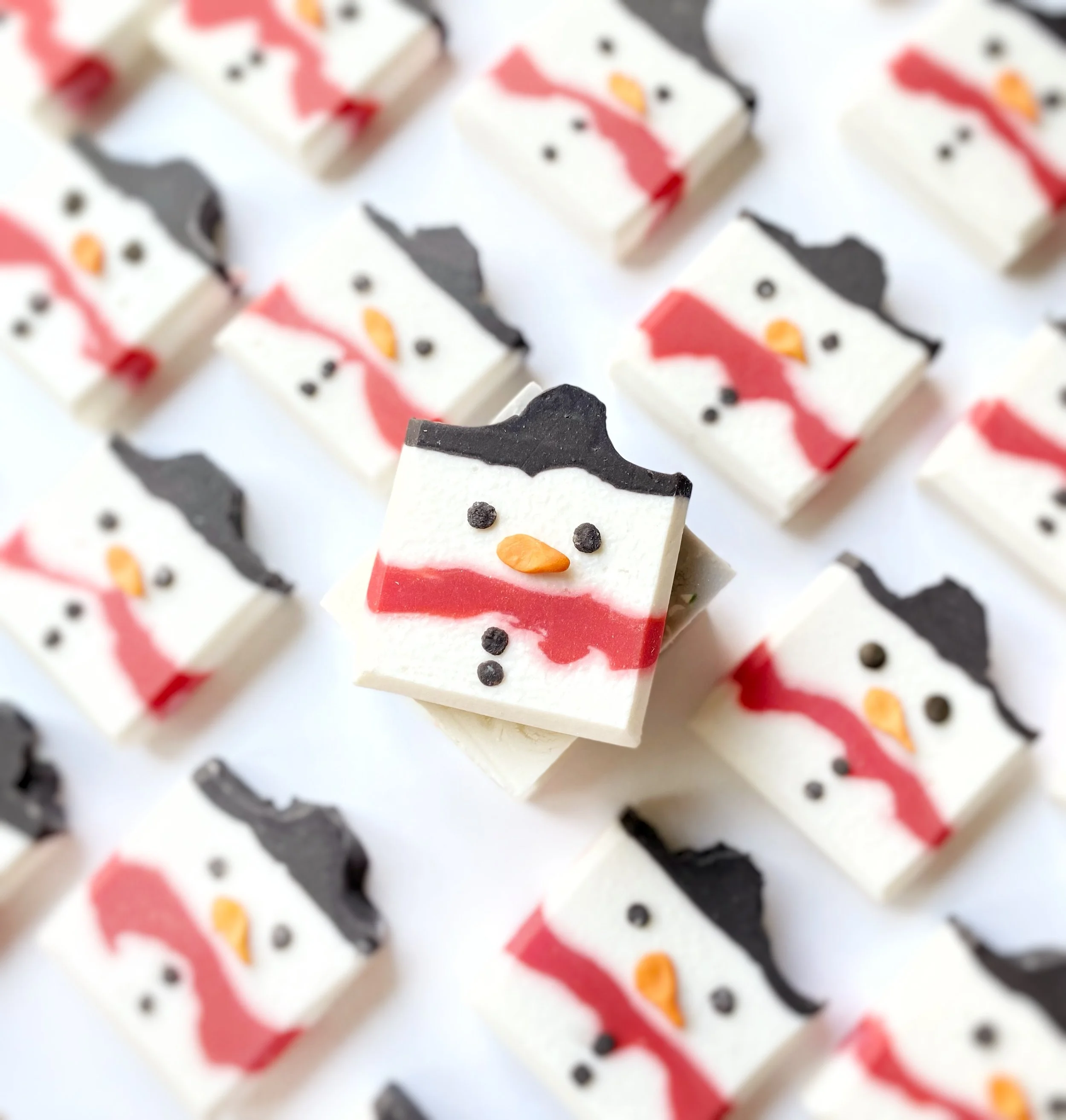 Assorted snowman-shaped marshmallows with black hats, orange noses, and red scarves, arranged on a white surface.