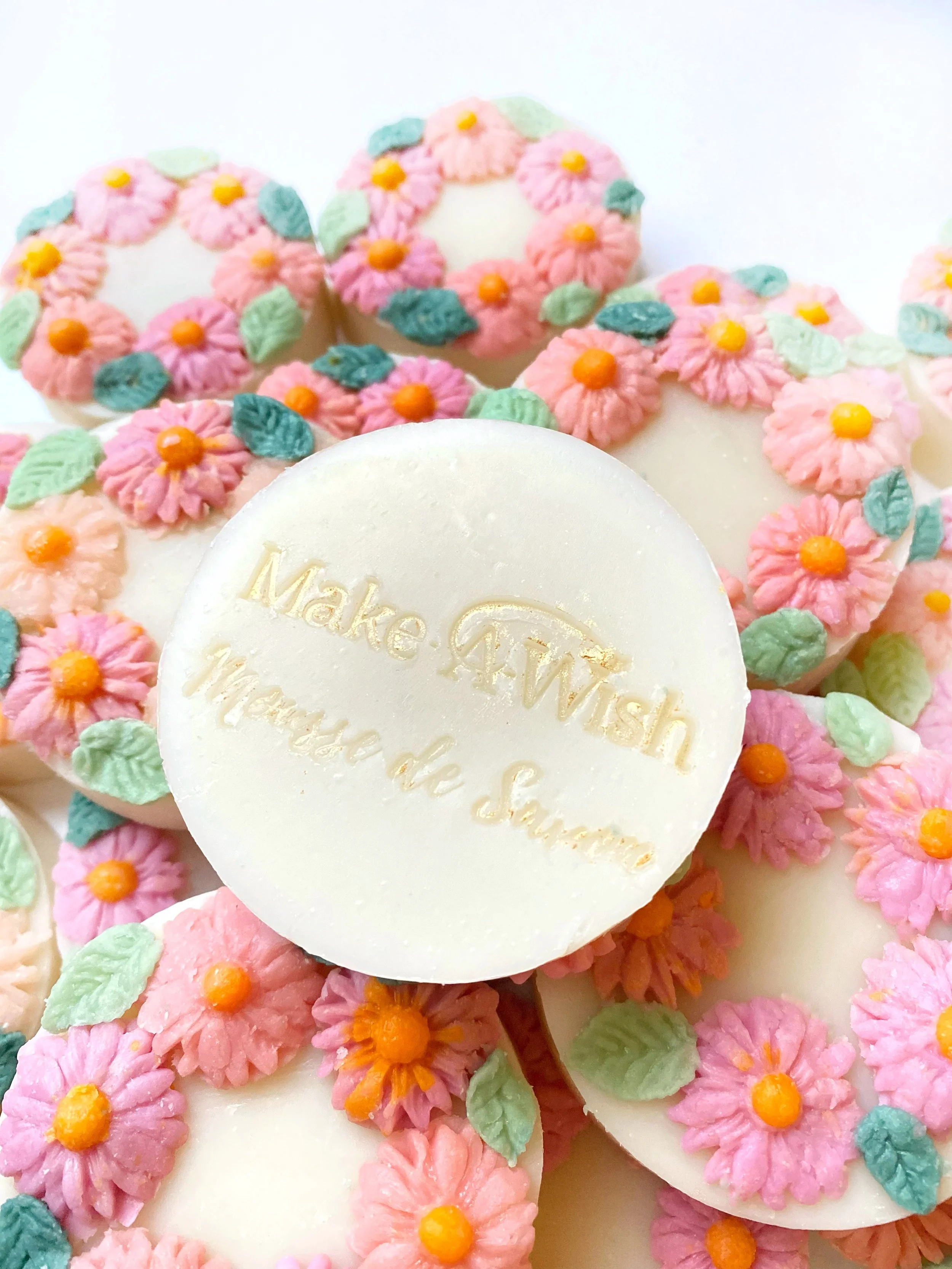White circular chocolate with the message 'Make A Wish I hope the Same' written on it, surrounded by decorated cookies with pink, yellow, and green frosting flowers and leaves.