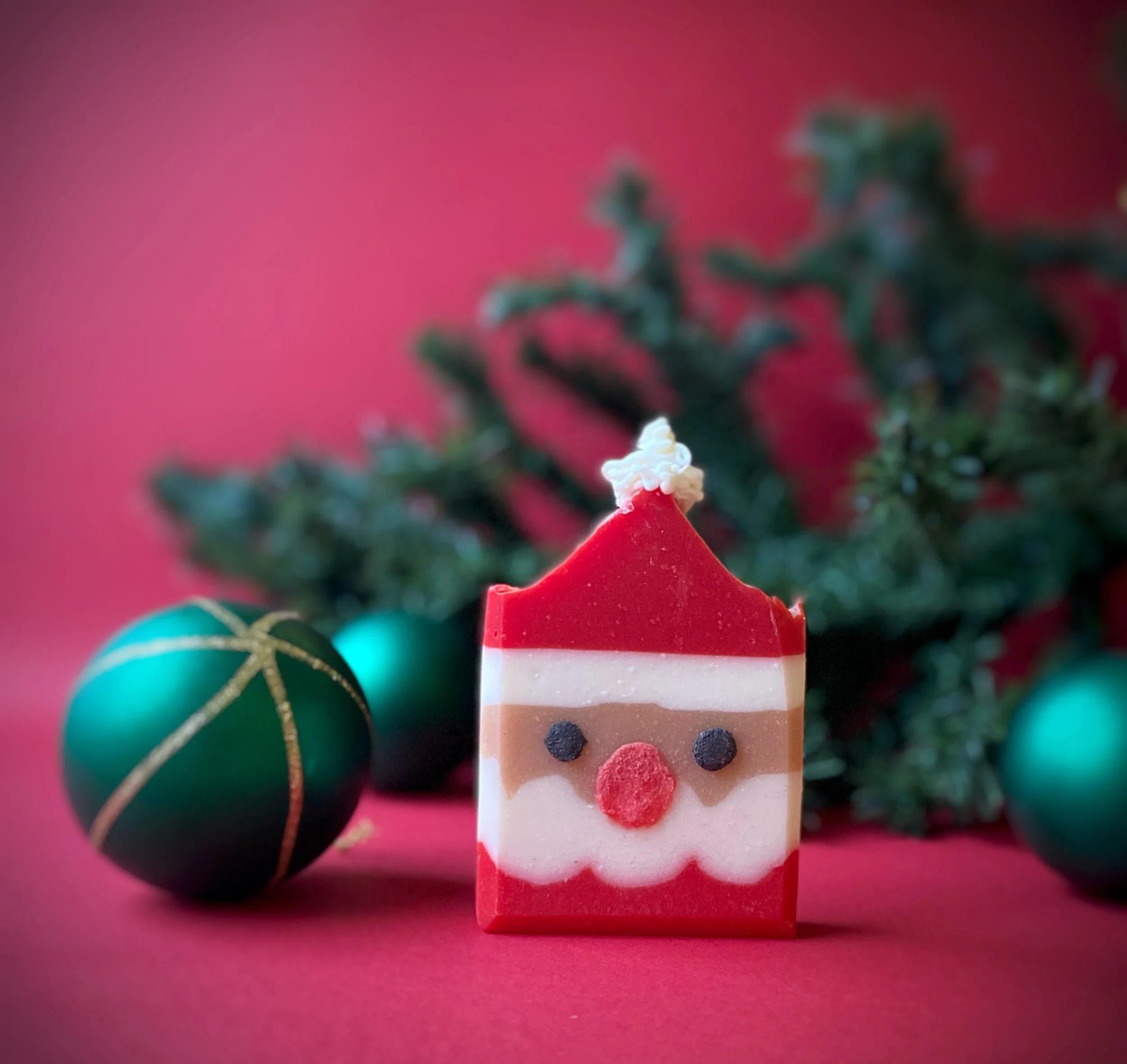 A Christmas-themed soap with Santa Claus face, surrounded by Christmas ornaments and a Christmas tree branch against a red background.