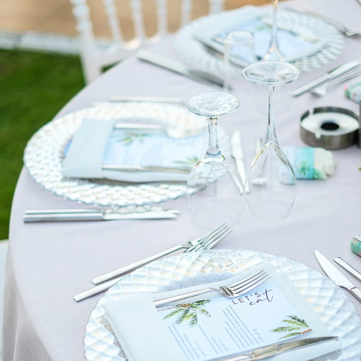 Table setting with white tablecloth, textured silver chargers, clear wine glasses, silverware, and printed menus. The menu has a palm tree design and says 'Let's Eat.' A small floral napkin is under the silverware.