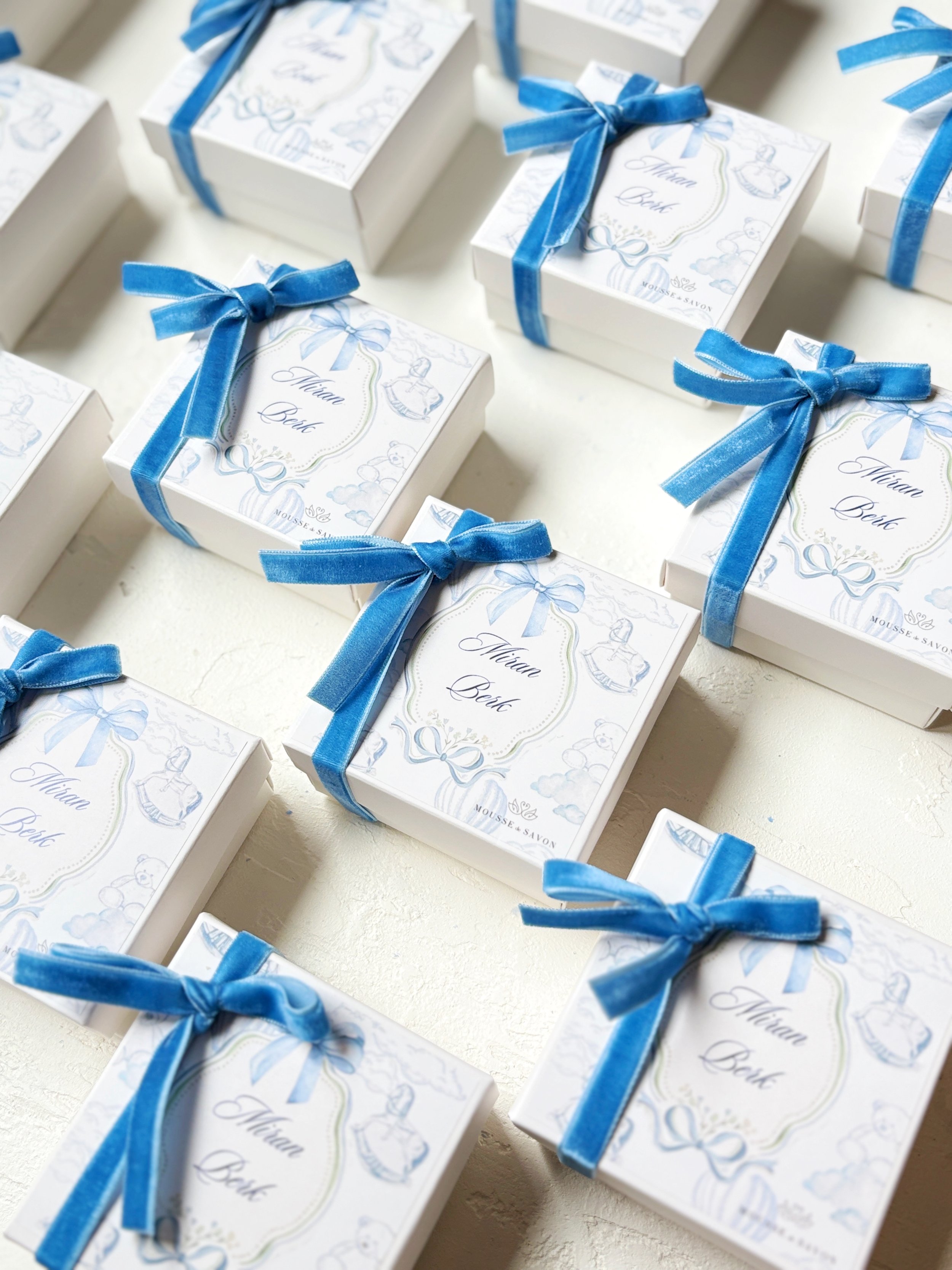 Multiple white gift boxes with blue ribbons and labels reading 'Miriam Beth' on a white surface.