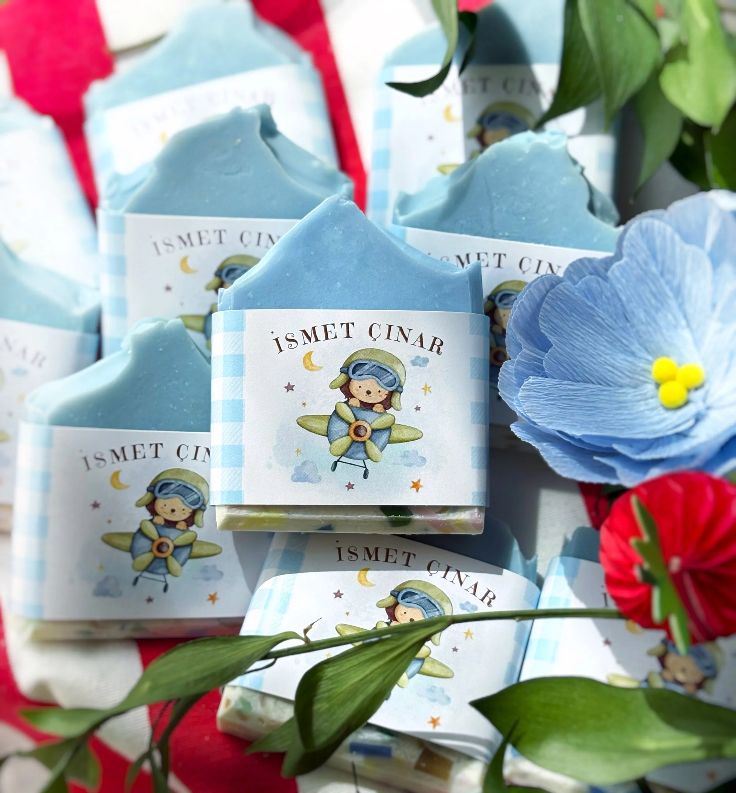 Several light blue soap bars with a label featuring a cartoon girl in a pilot outfit, flying an airplane. The soaps are placed among red and blue paper flowers and green leaves, with a red cloth in the background.