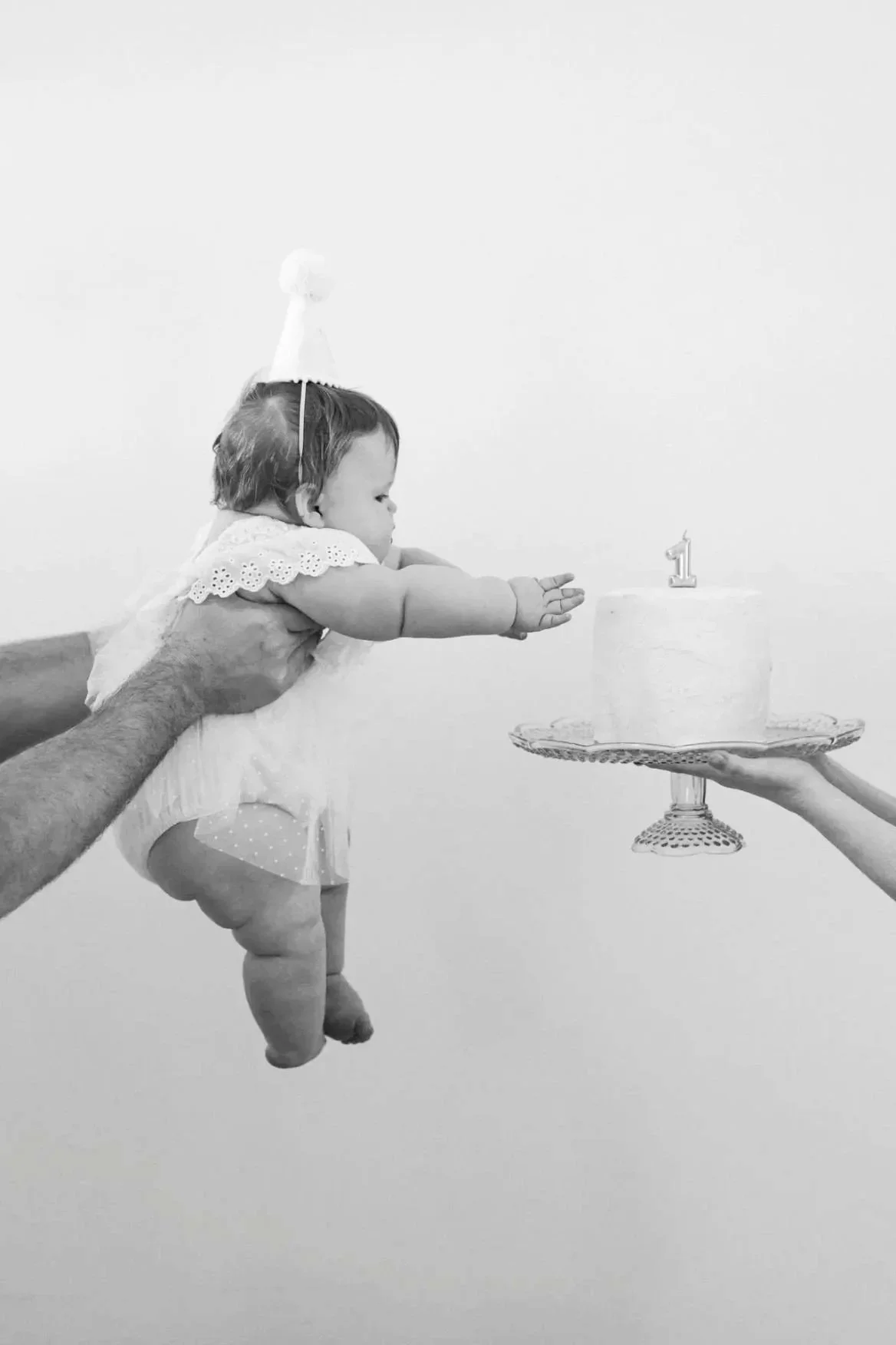 A baby being held horizontally with arms outstretched towards a birthday cake with a number one candle, in a celebration setting.