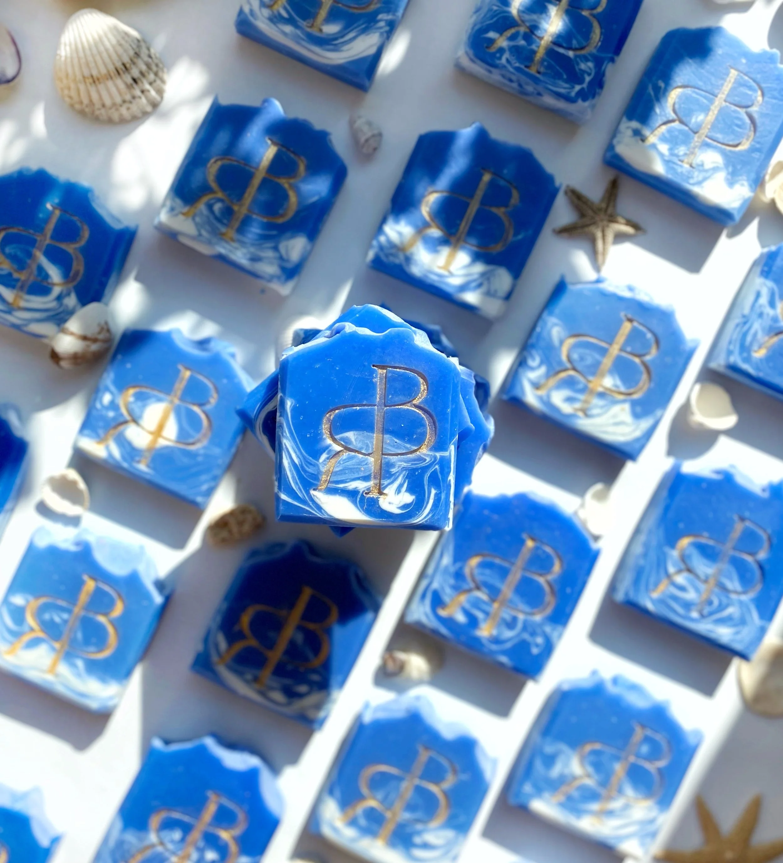 Several blue and white marbled soap bars with a gold symbol featuring a 'B' and a vertical line through it, surrounded by small seashells and starfish on a white surface.