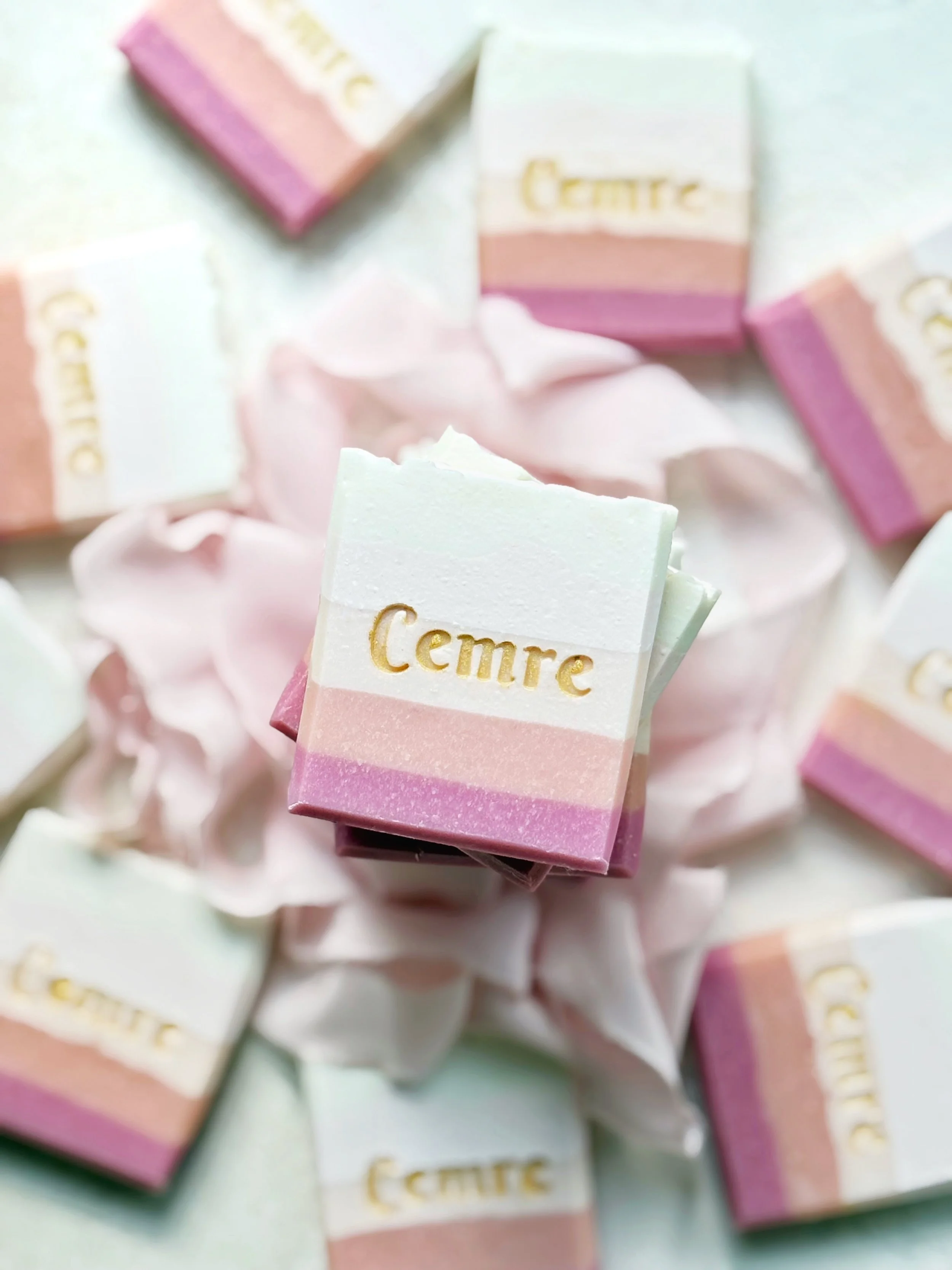 Colorful layered soap bars with the word "Cemre" embossed in gold on the top layer, surrounded by pink tissue paper.