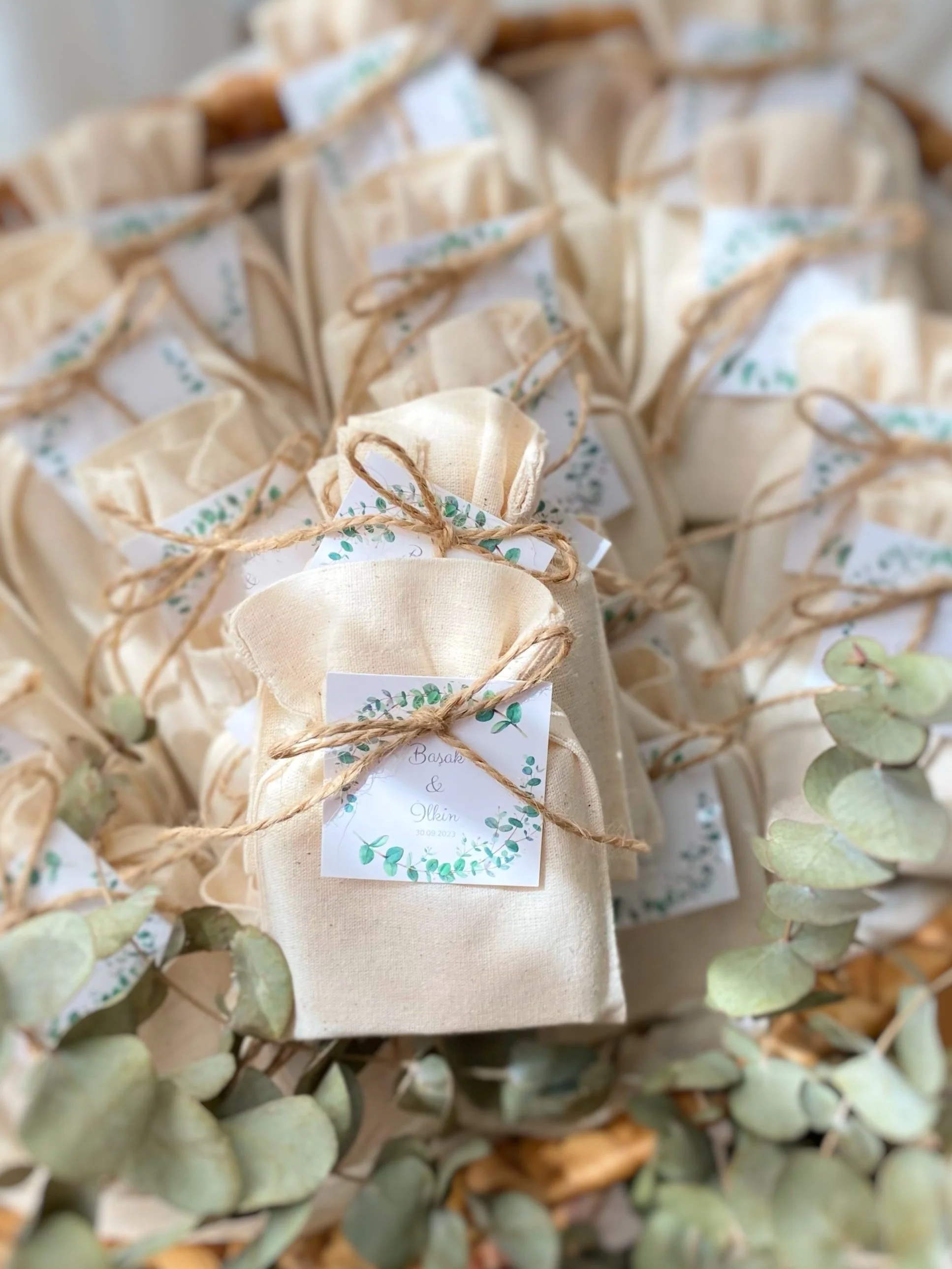 Multiple beige fabric gift bags tied with twine, each with a small decorative label, surrounded by eucalyptus leaves.