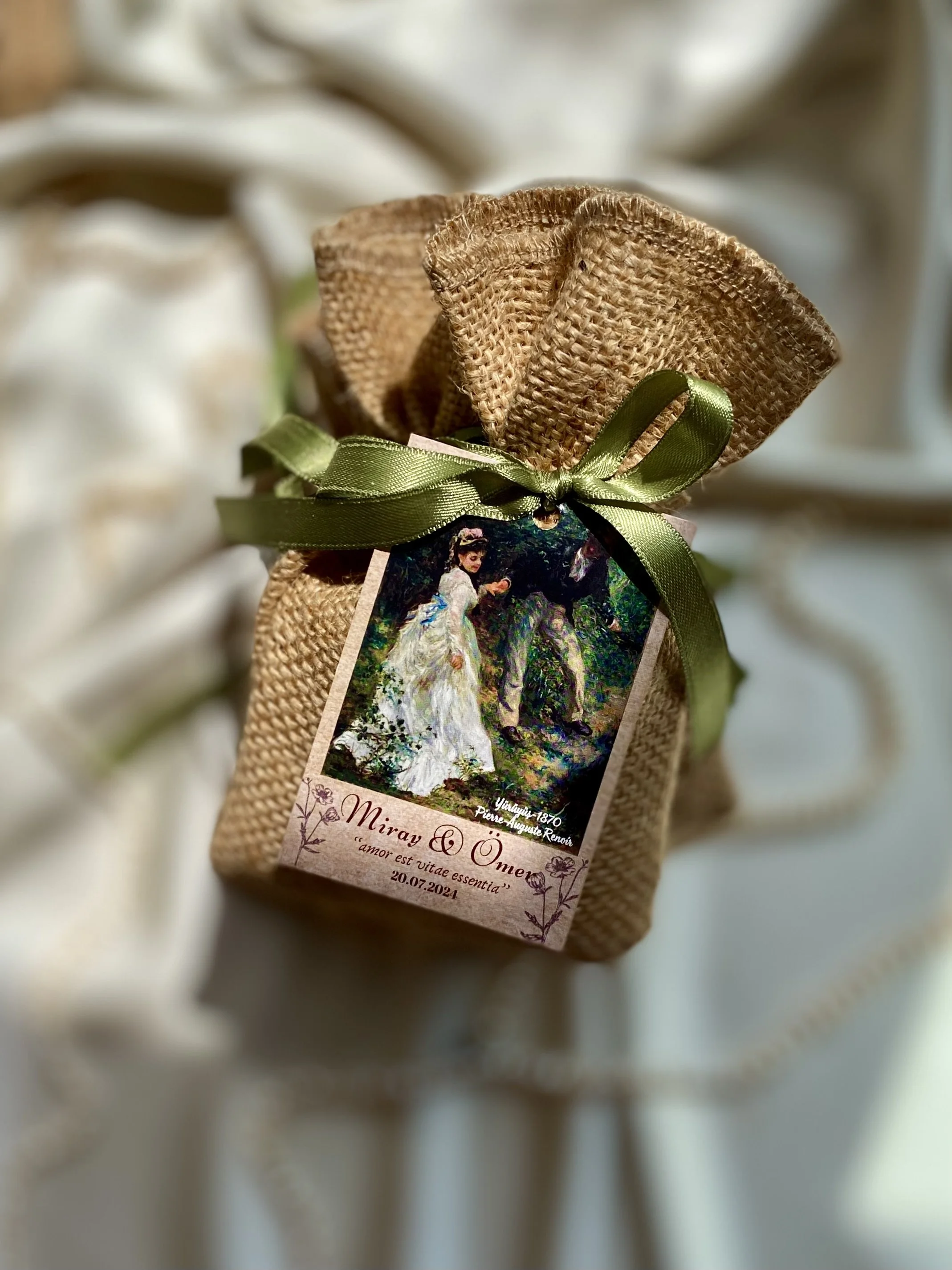 A small gift wrapped in burlap with a green ribbon bow, attached with a tag featuring a painting of a woman in a white dress and text in Latin.