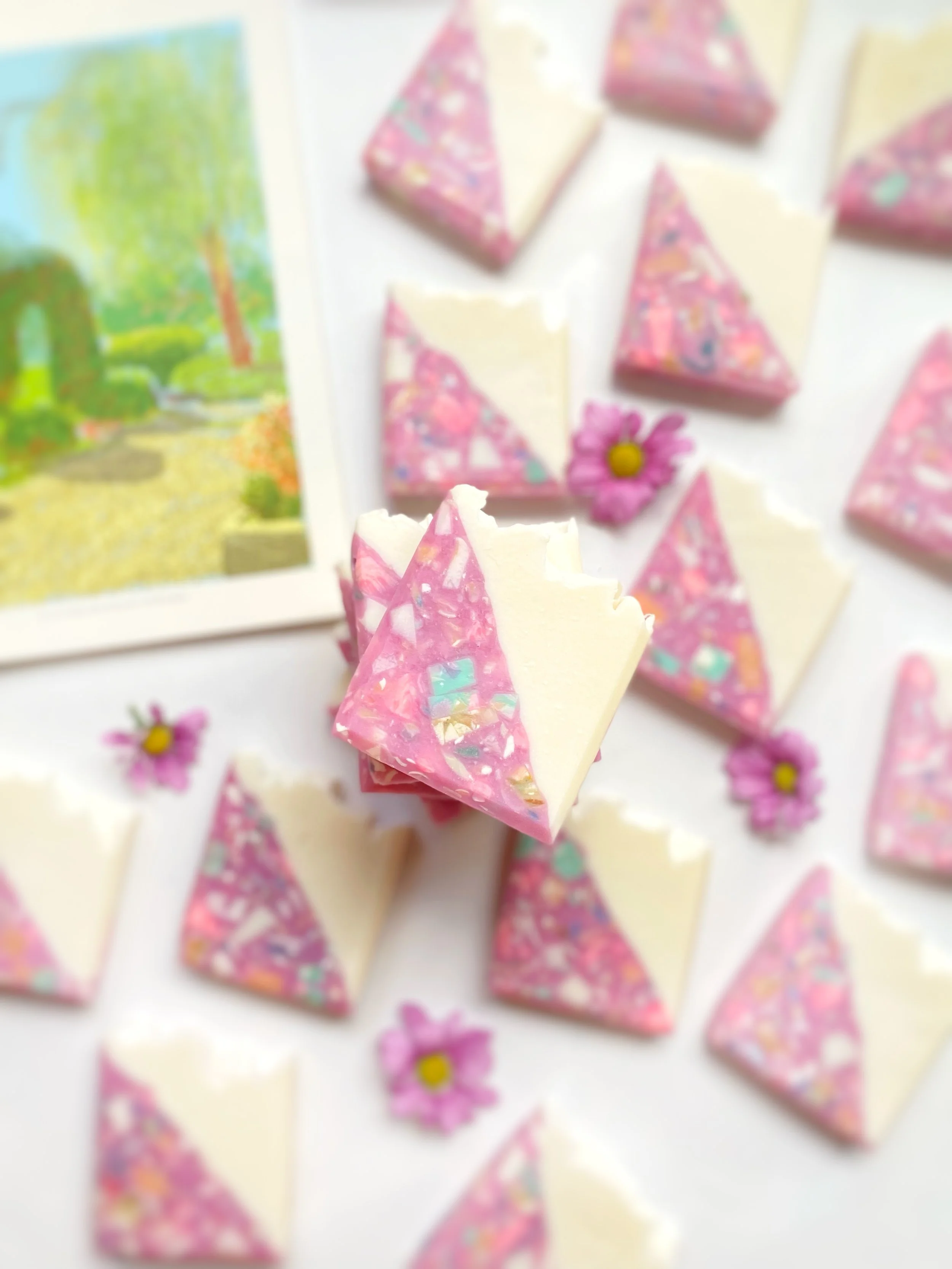 Assorted pink and white chocolate bark pieces with floral and abstract designs, garnished with small pink flowers, on a white surface with a colorful painting in the background.